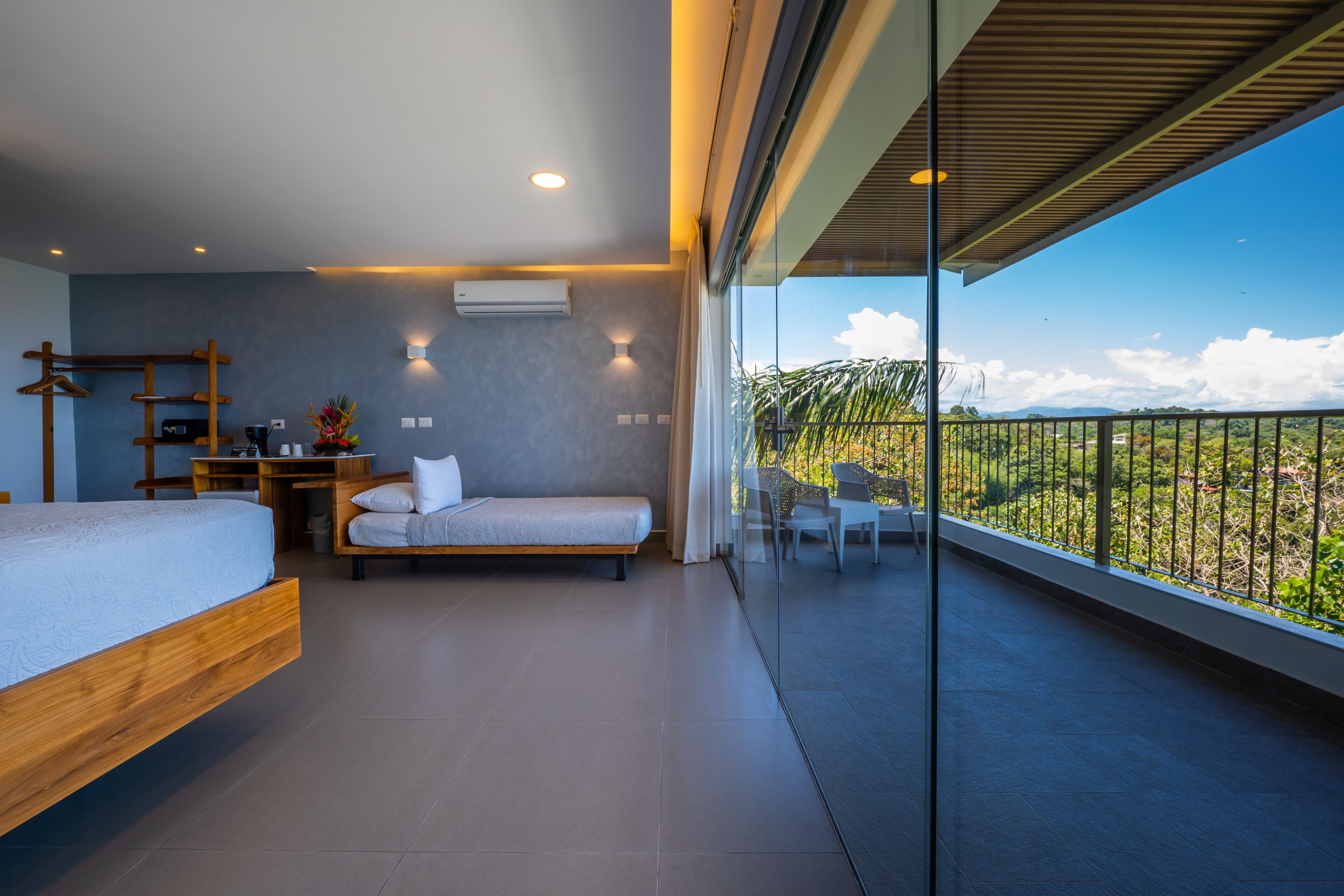 Un dormitorio moderno con cama, mesita de noche y escritorio con silla. Hay un balcón con vistas a zonas verdes y montañas.