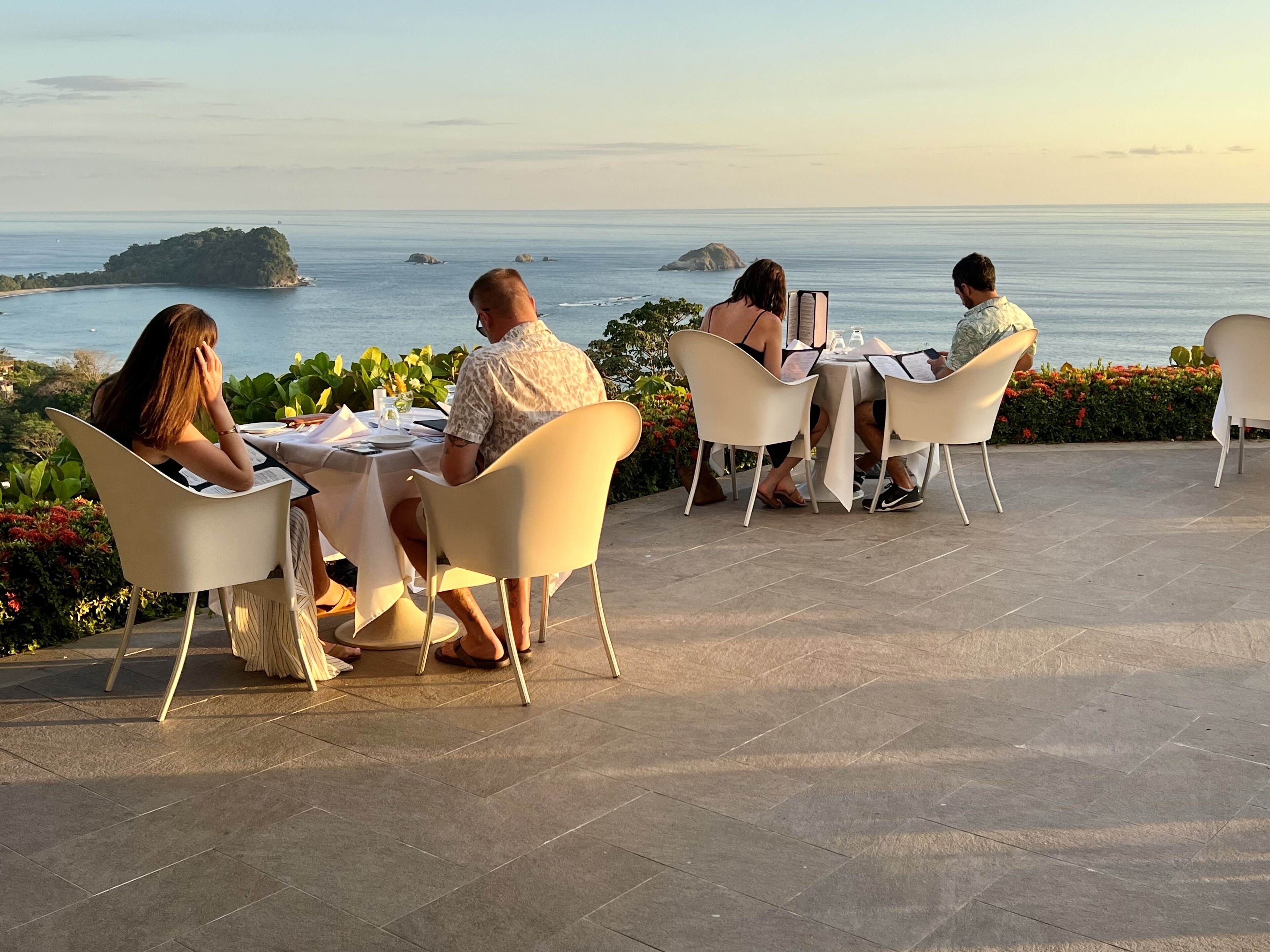 A group of people dining at an outdoor table with a view of the sea and an island.