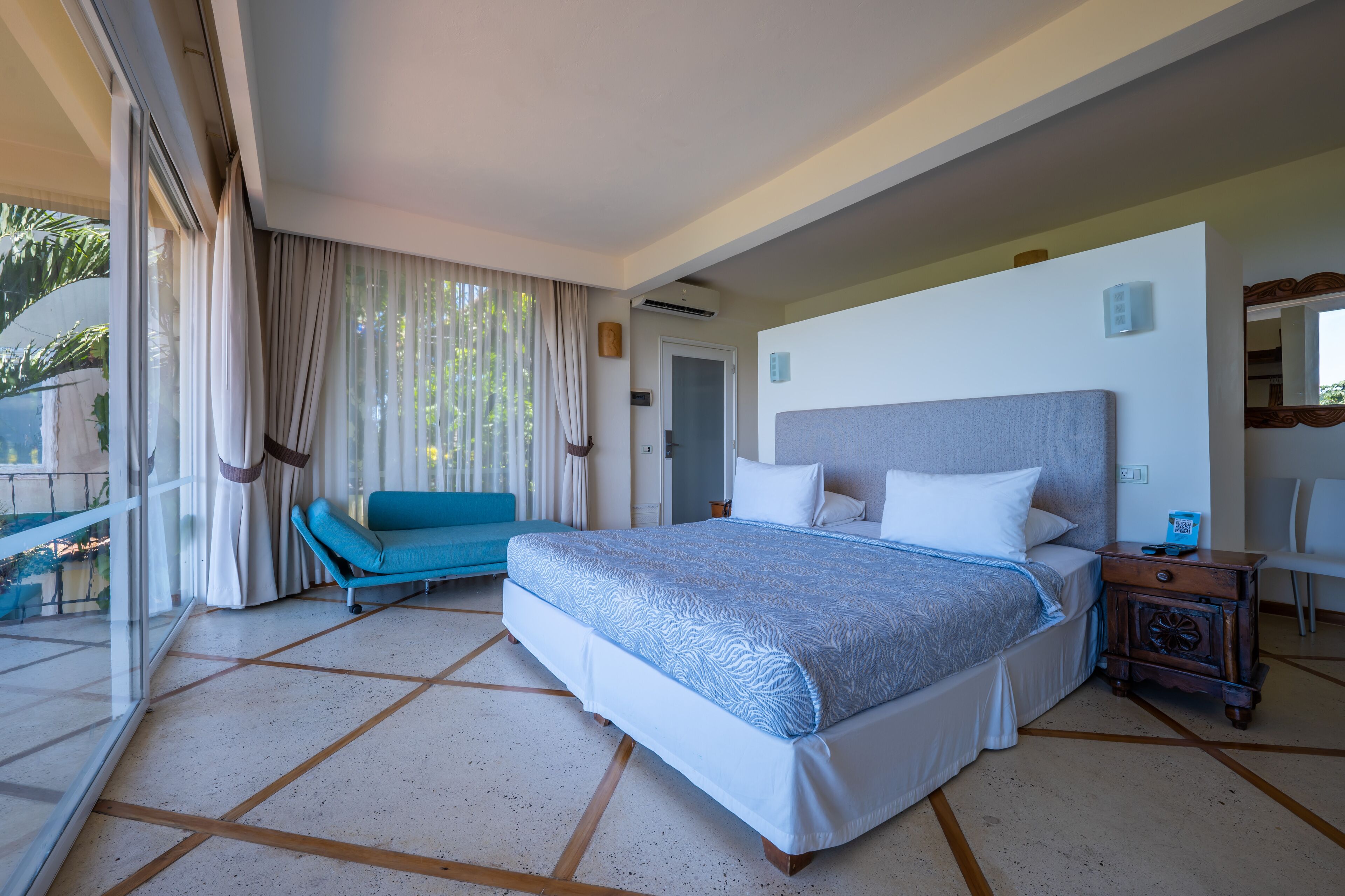 A bedroom with a large bed, a blue sofa, a balcony with curtains, and a small table with a lamp.