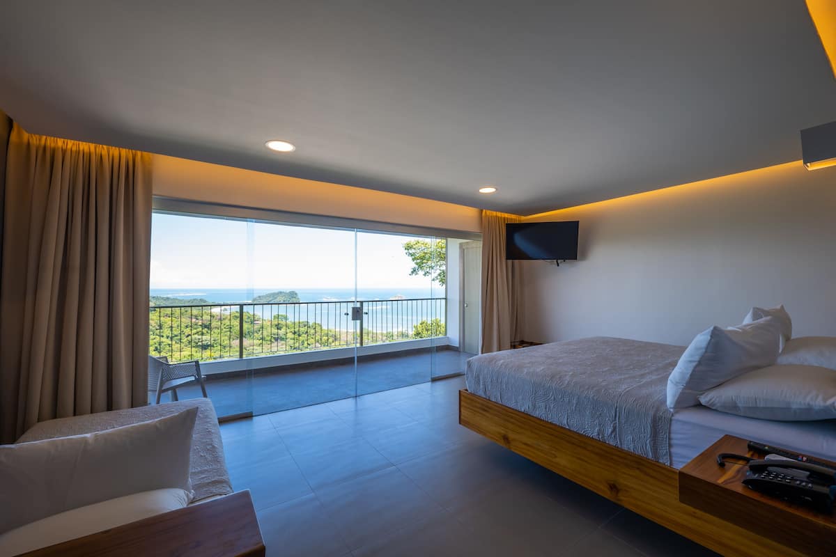 Une chambre moderne avec un grand lit, une table de chevet avec un téléphone, un téléviseur fixé au mur et un balcon offrant une vue sur le paysage.