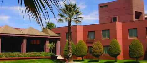 A swimming pool with lounge chairs and a palm tree, surrounded by a building with a red facade and a clear blue sky.