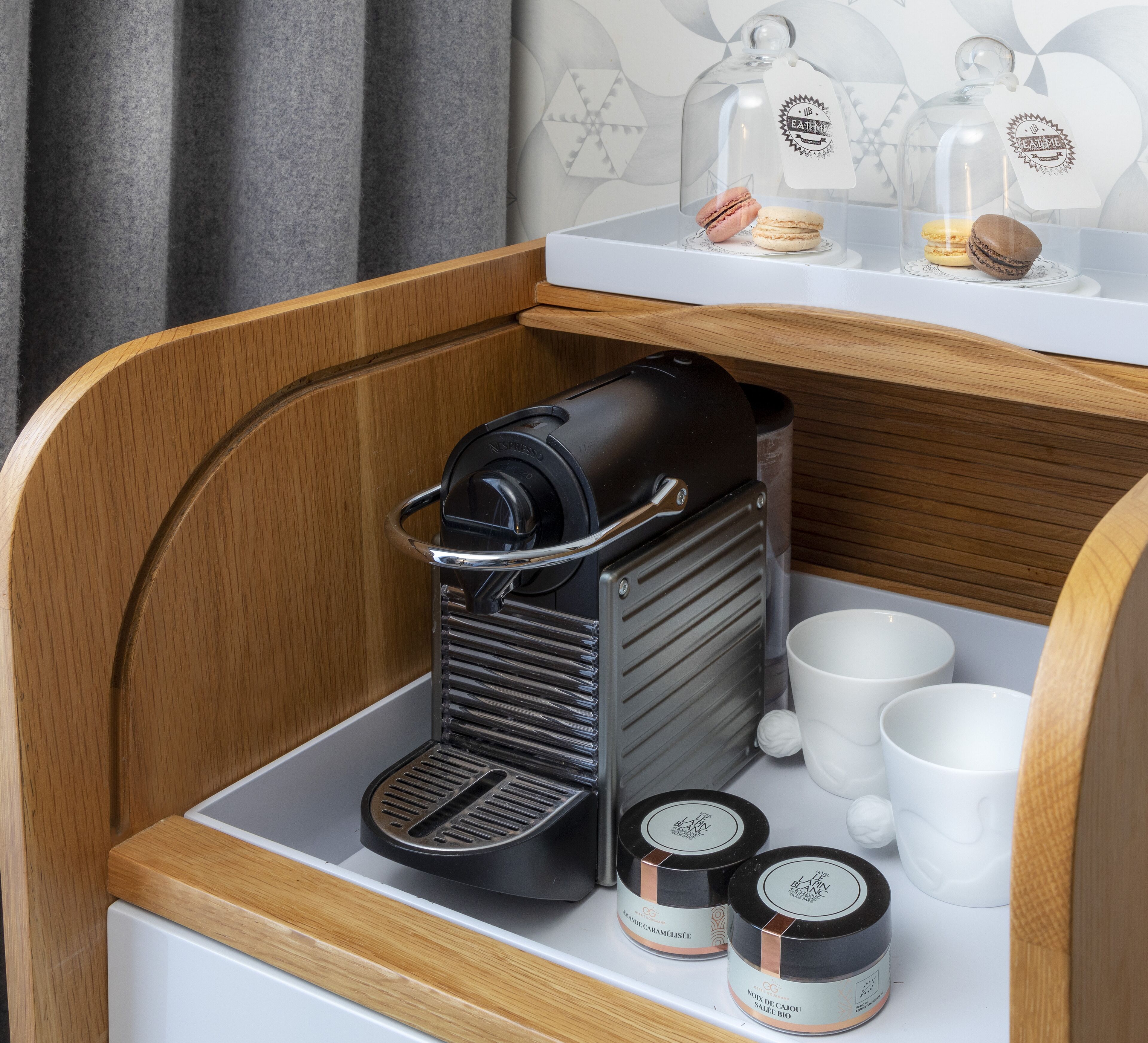 Une petite machine à café, placée dans un meuble en bois, accompagnée de deux tasses blanches et de deux bocaux contenant des aliments.