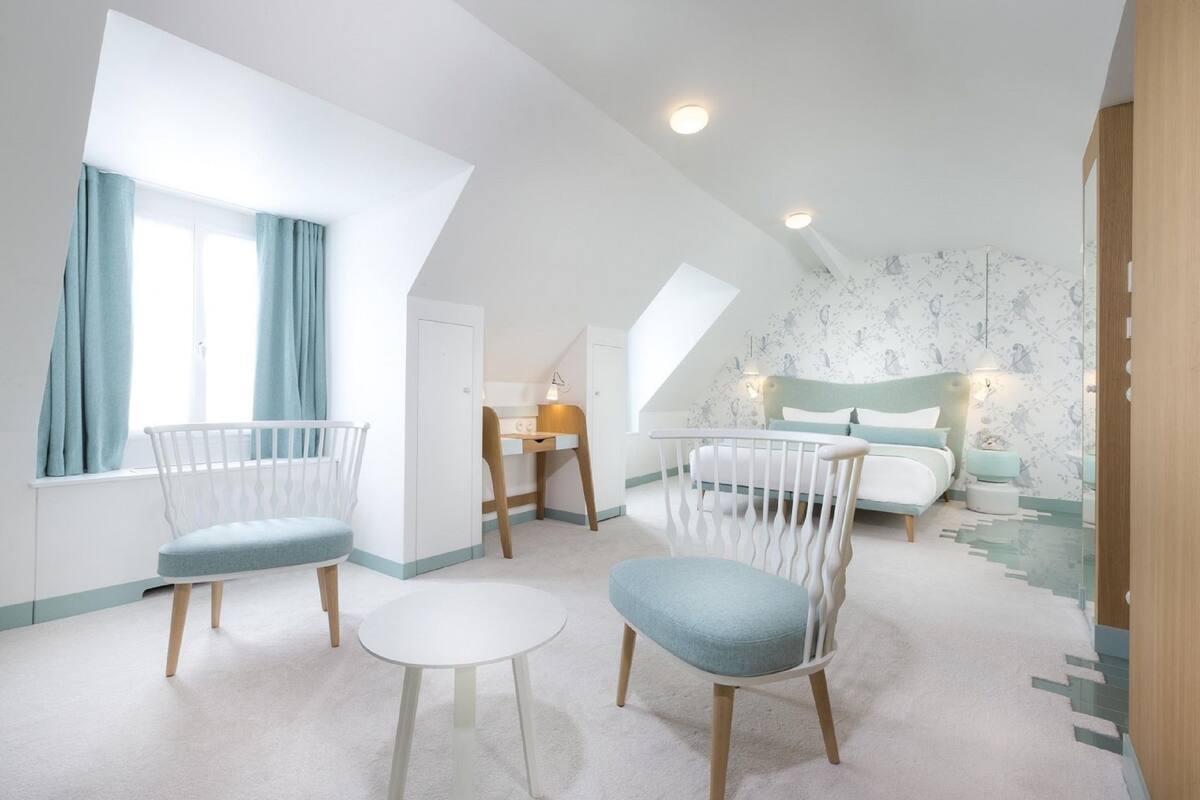 A spacious room with a high ceiling, a large window with curtains, a white bed, a white chair, and a small table.
