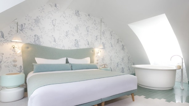 A bedroom with a bed, a nightstand, a lamp, a wall-mounted light, and a bathtub. - Le Lapin Blanc (Paris)