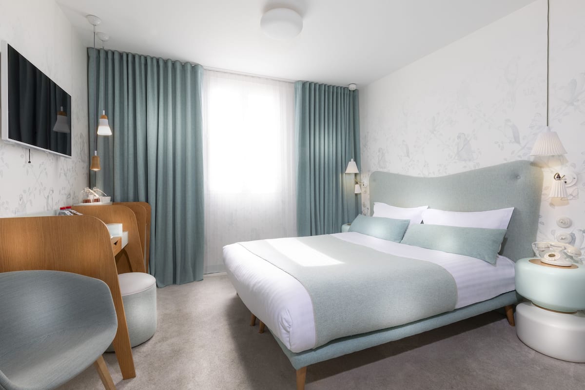 A modern hotel room with a bed, a desk, a chair, and a nightstand.