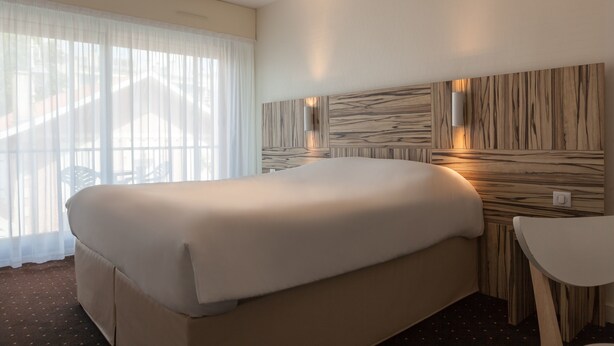 Quarto de hotel com uma cama grande, uma mesinha e um candeeiro fixado na parede.