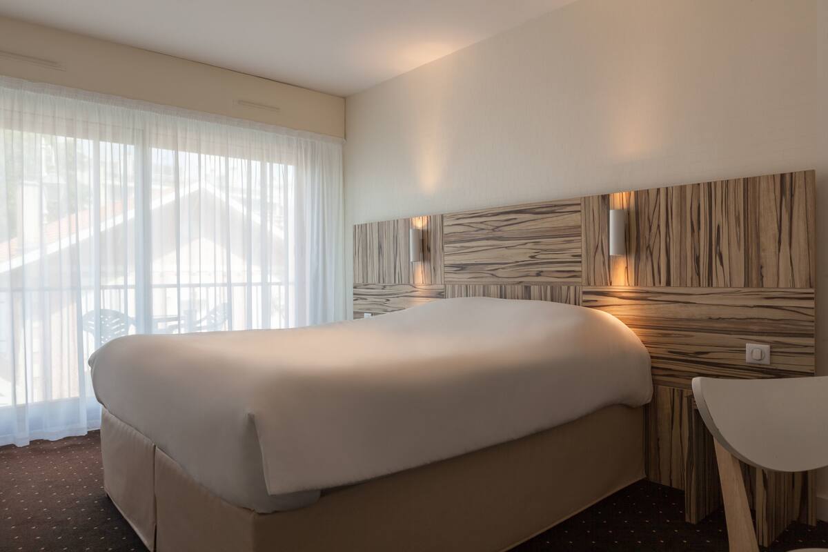 A hotel room with a large bed, a small table, and a wall-mounted light fixture.