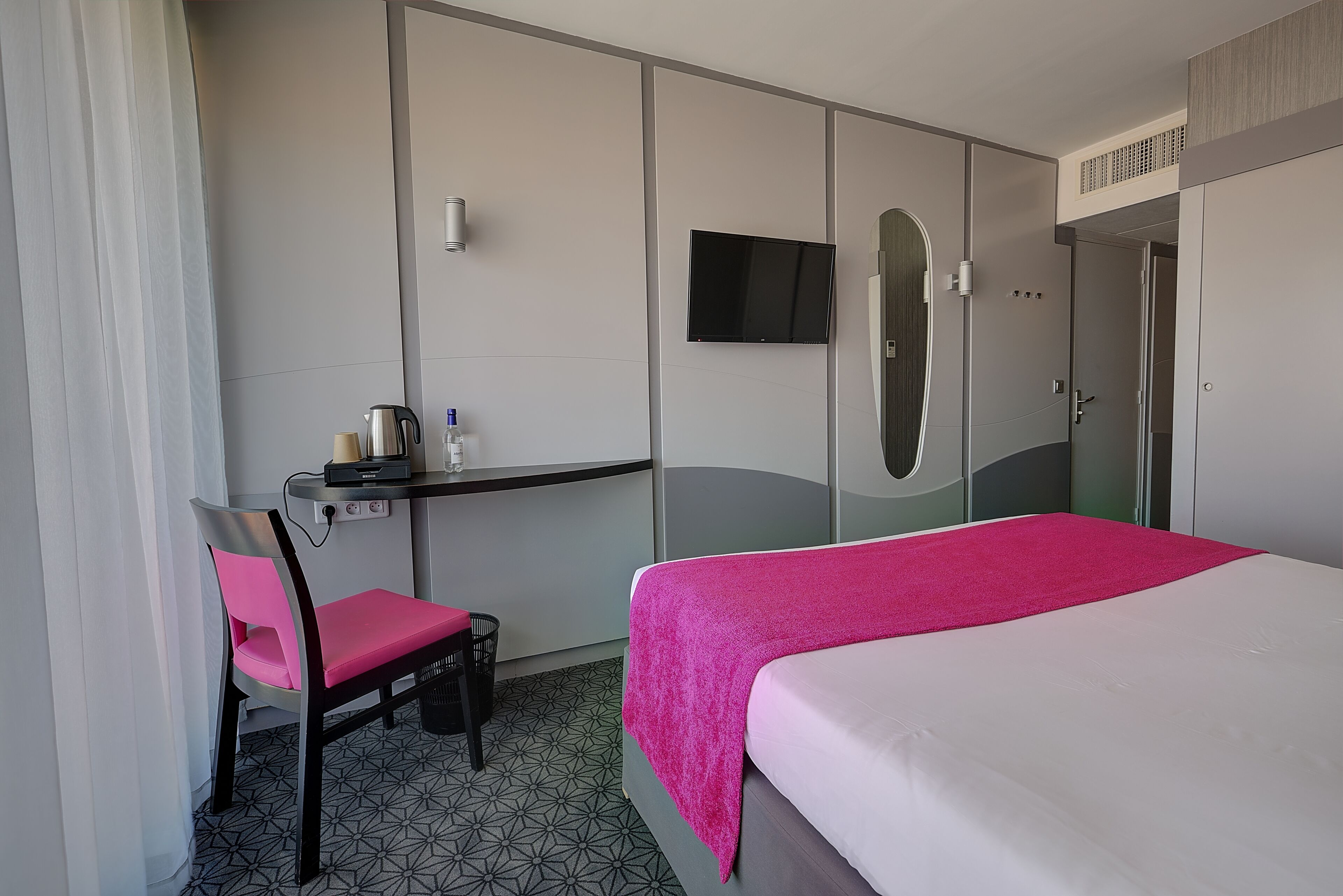 A hotel room with a bed, a desk with a chair, a TV mounted on the wall, and a wardrobe.