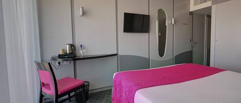 Una habitación de hotel con cama, escritorio con silla, televisor montado en la pared y armario.