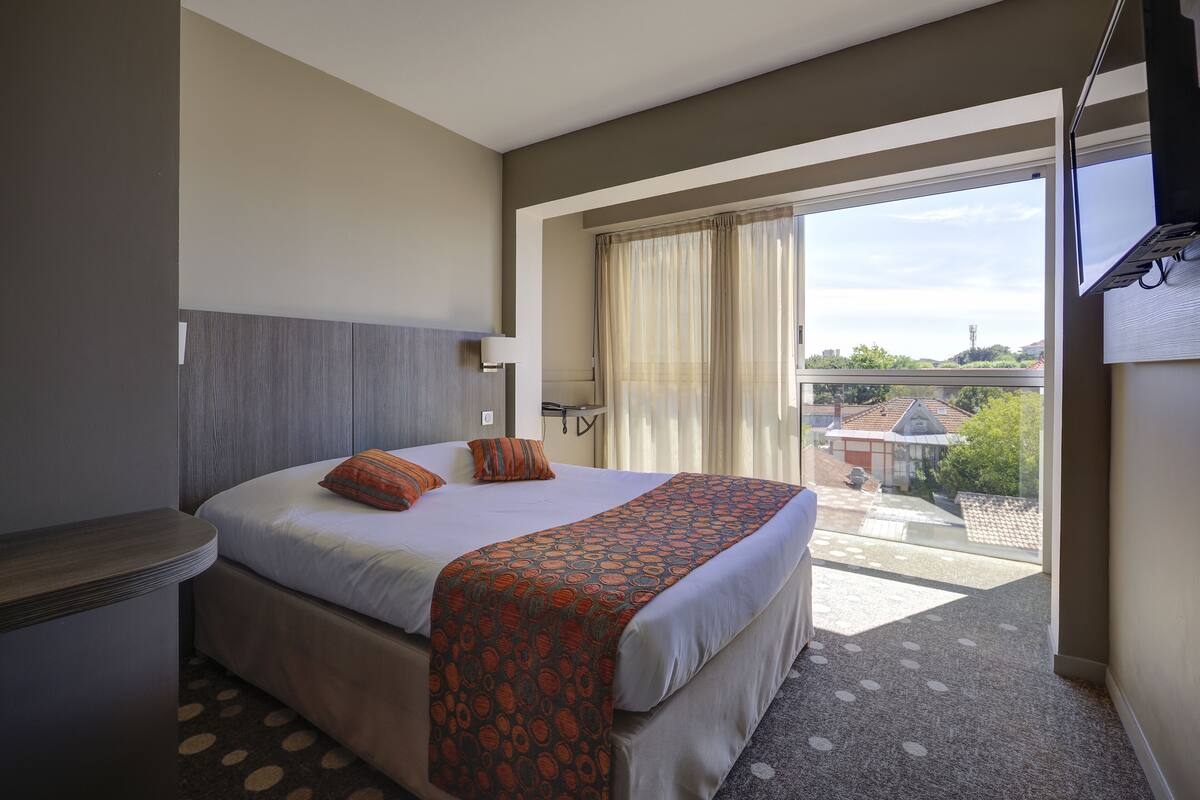 A hotel room with a large bed, a nightstand, a window with curtains, and a view of buildings outside.