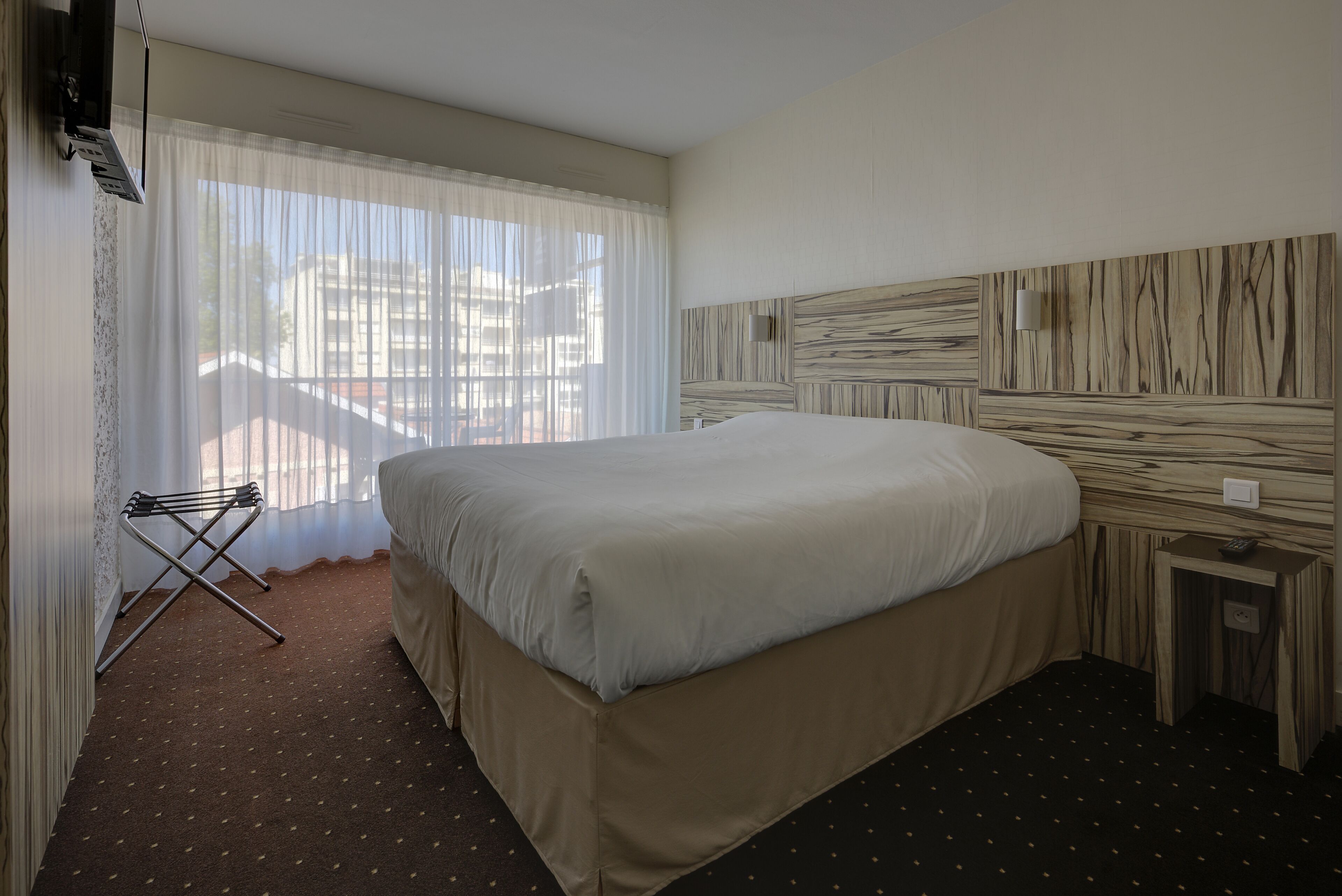 A hotel room with a large bed, a wooden headboard, a small bedside table, and a window with sheer curtains.