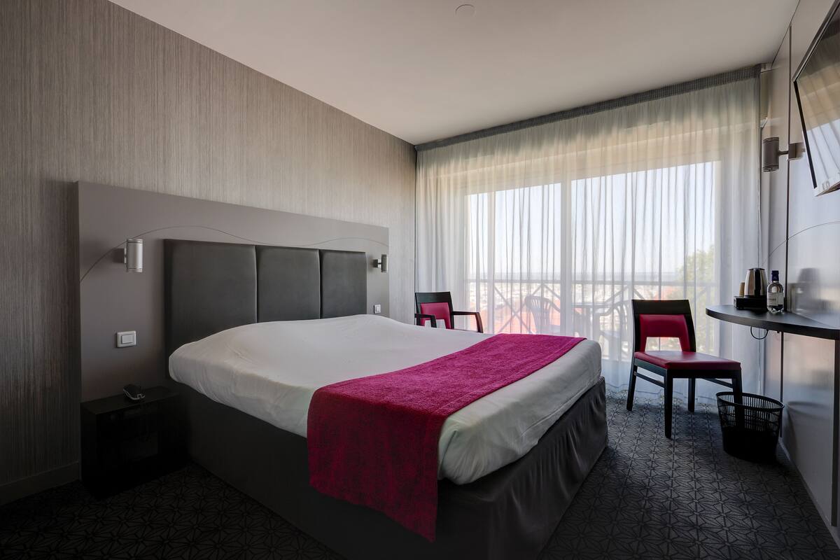 A modern hotel room with a large bed, a desk with a chair, and a balcony with a view.