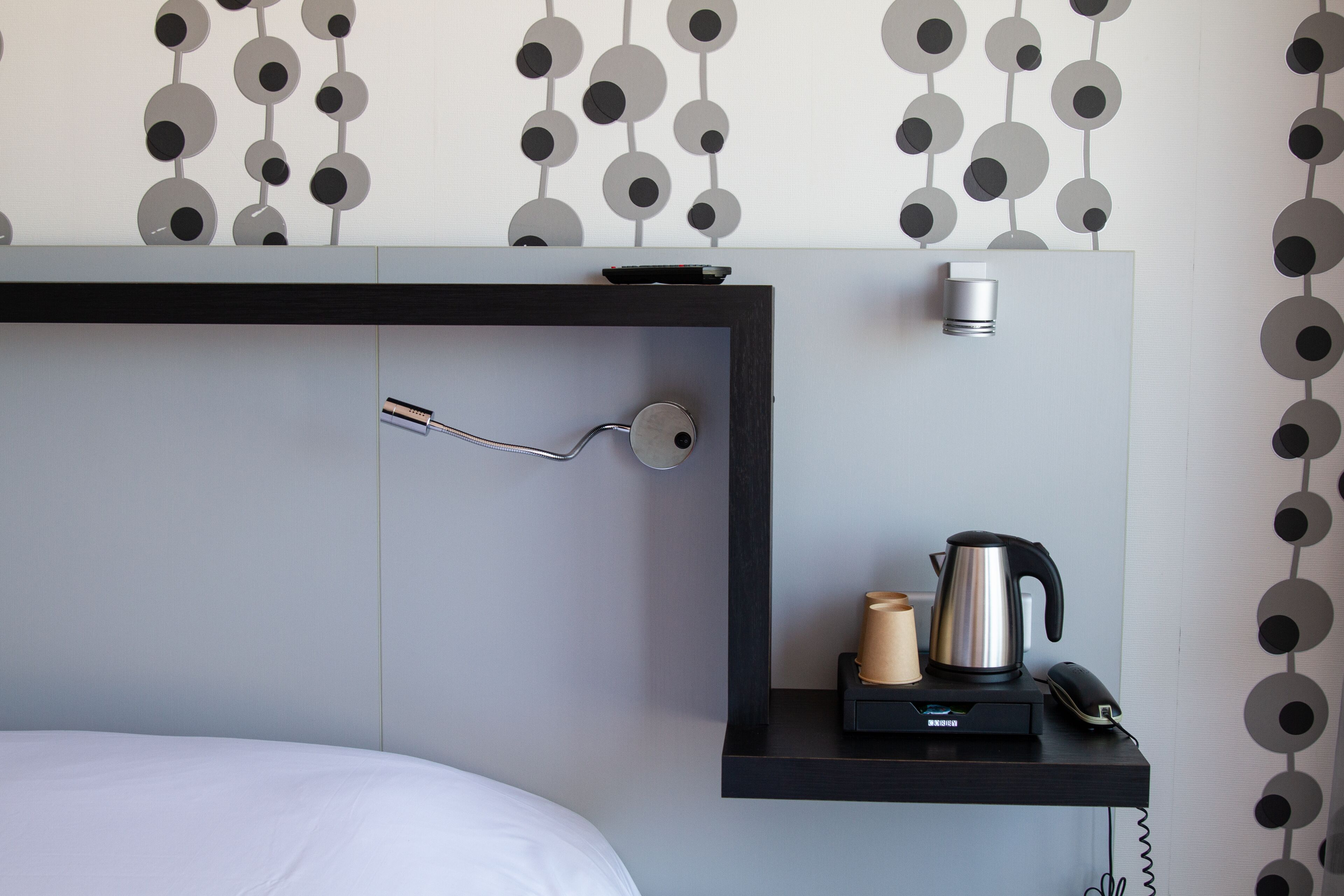 A modern hotel room with a bed, a bedside table with a kettle, a phone, and a small lamp.