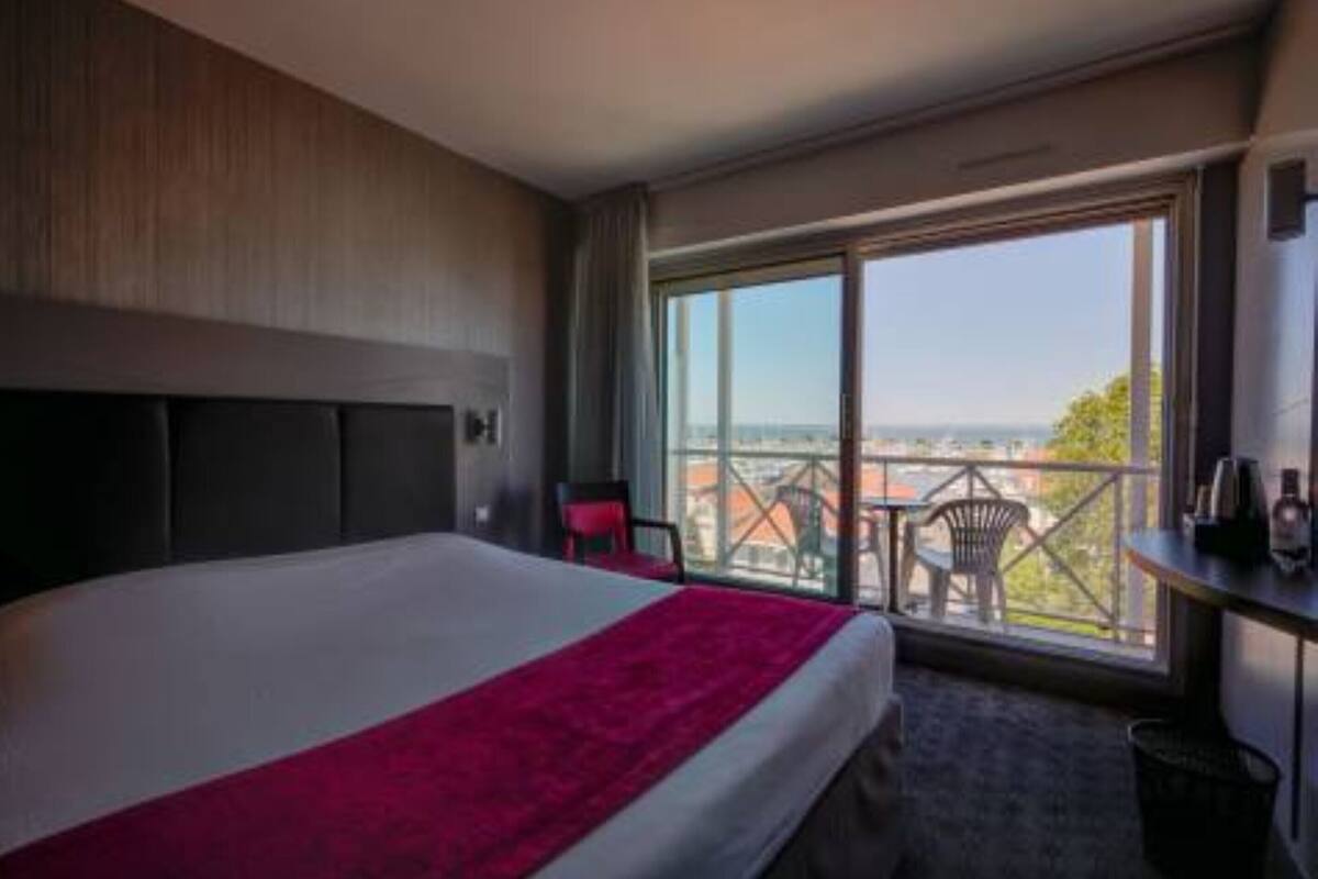 A hotel room with a large bed, a red throw blanket, a view of the city through a large window, and a balcony with chairs.