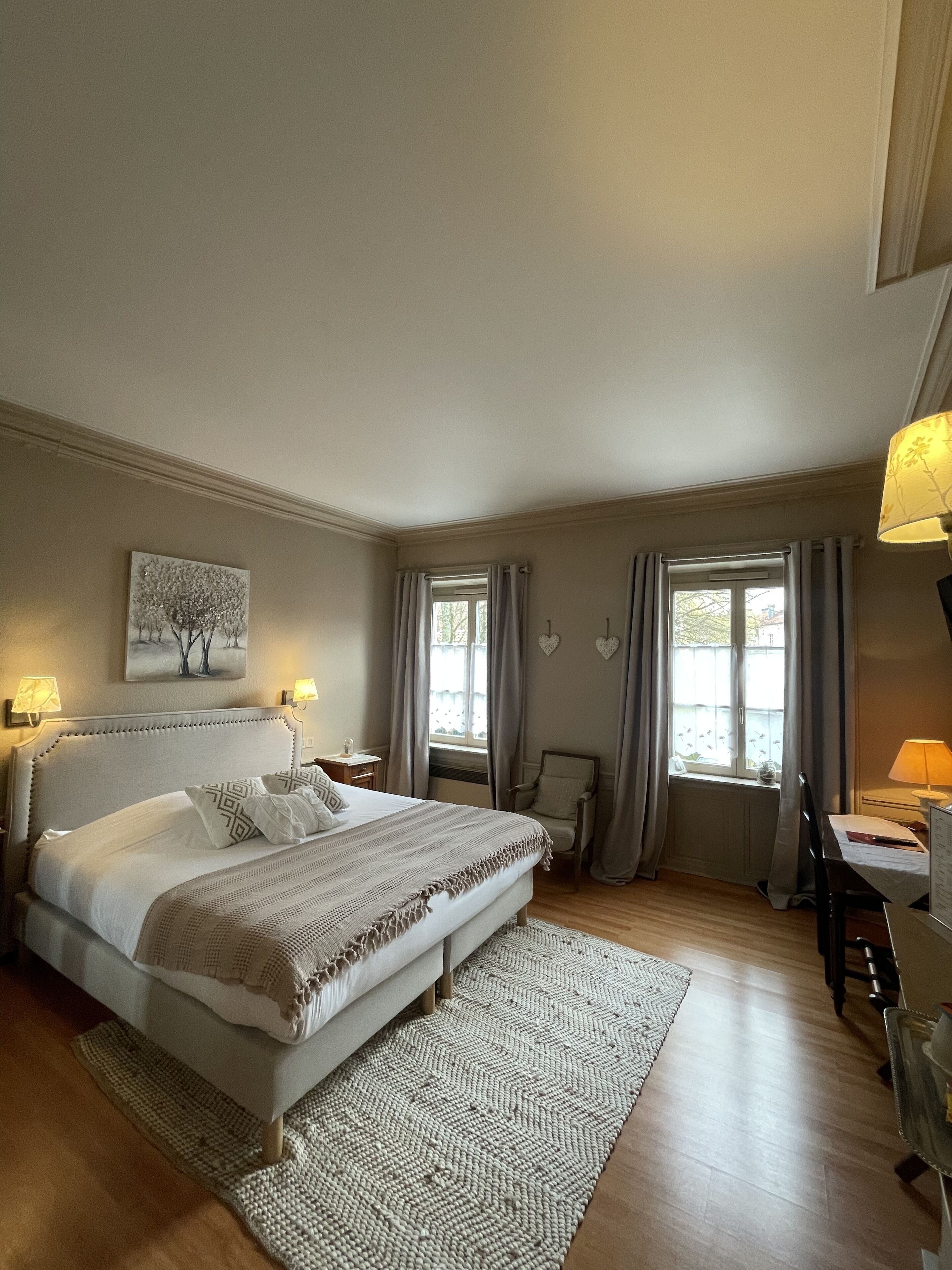 A bedroom with a large bed, a desk, two windows with curtains, a chair, and a painting on the wall.