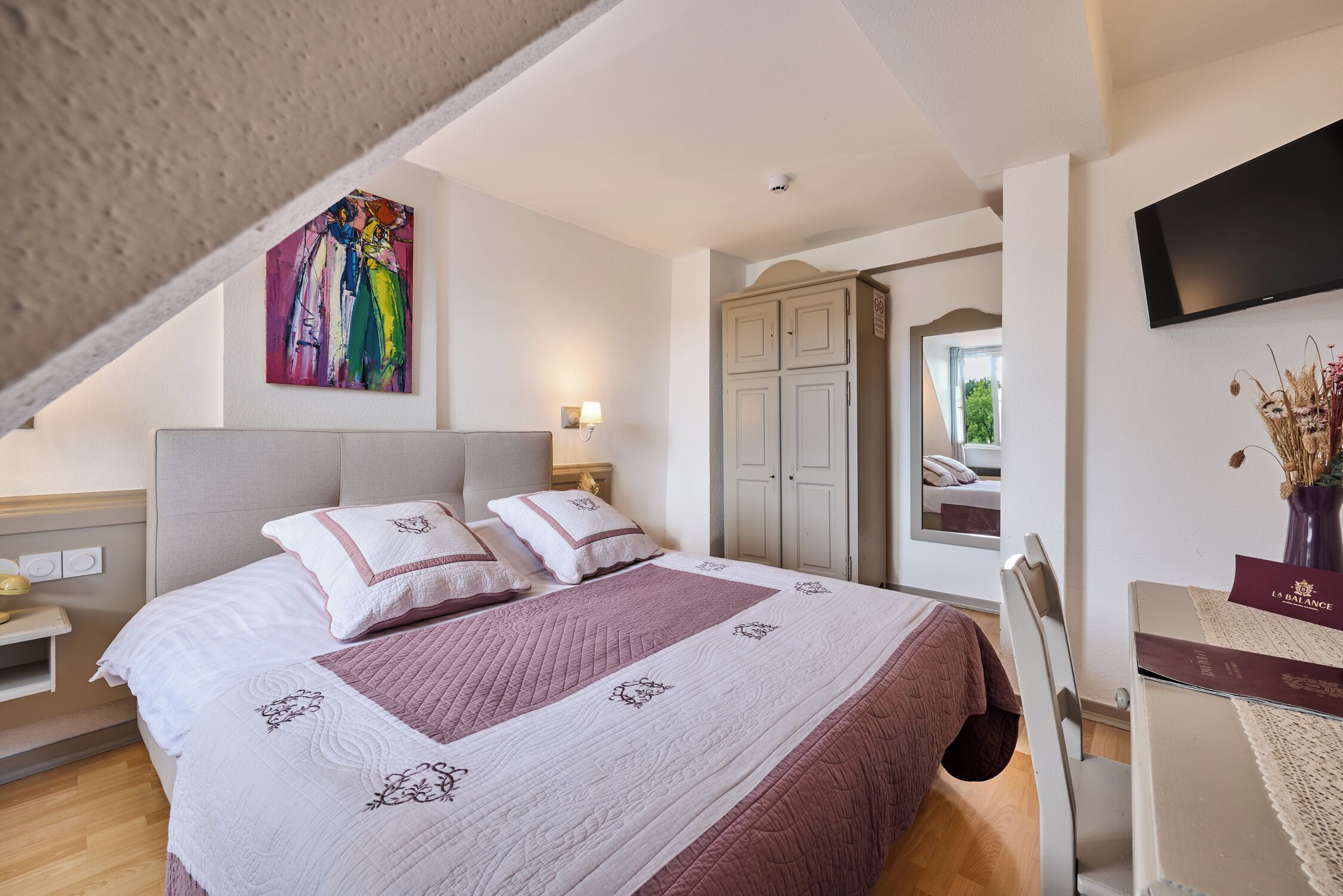 A bedroom with a bed, a painting, a wardrobe, a TV, and a desk with a vase.