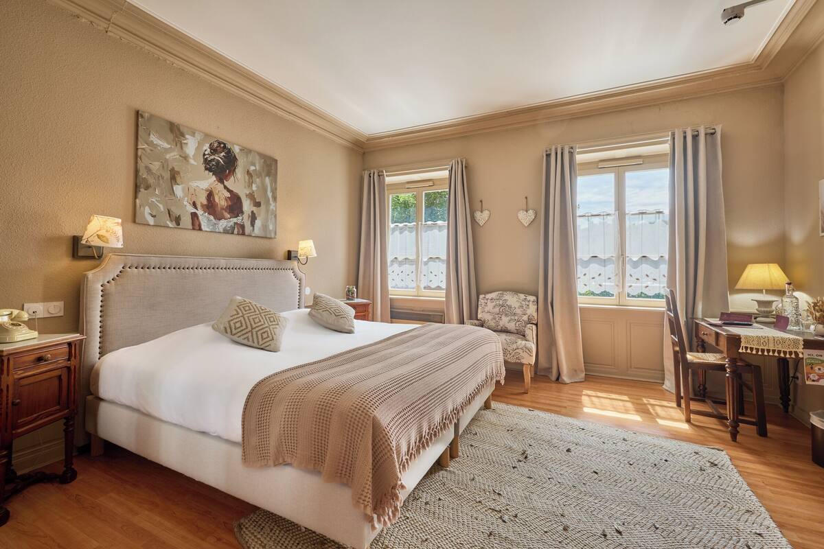 Een slaapkamer met een groot bed, een nachtkastje met lamp, een stoel, een bureau met lamp en een raam met gordijnen.