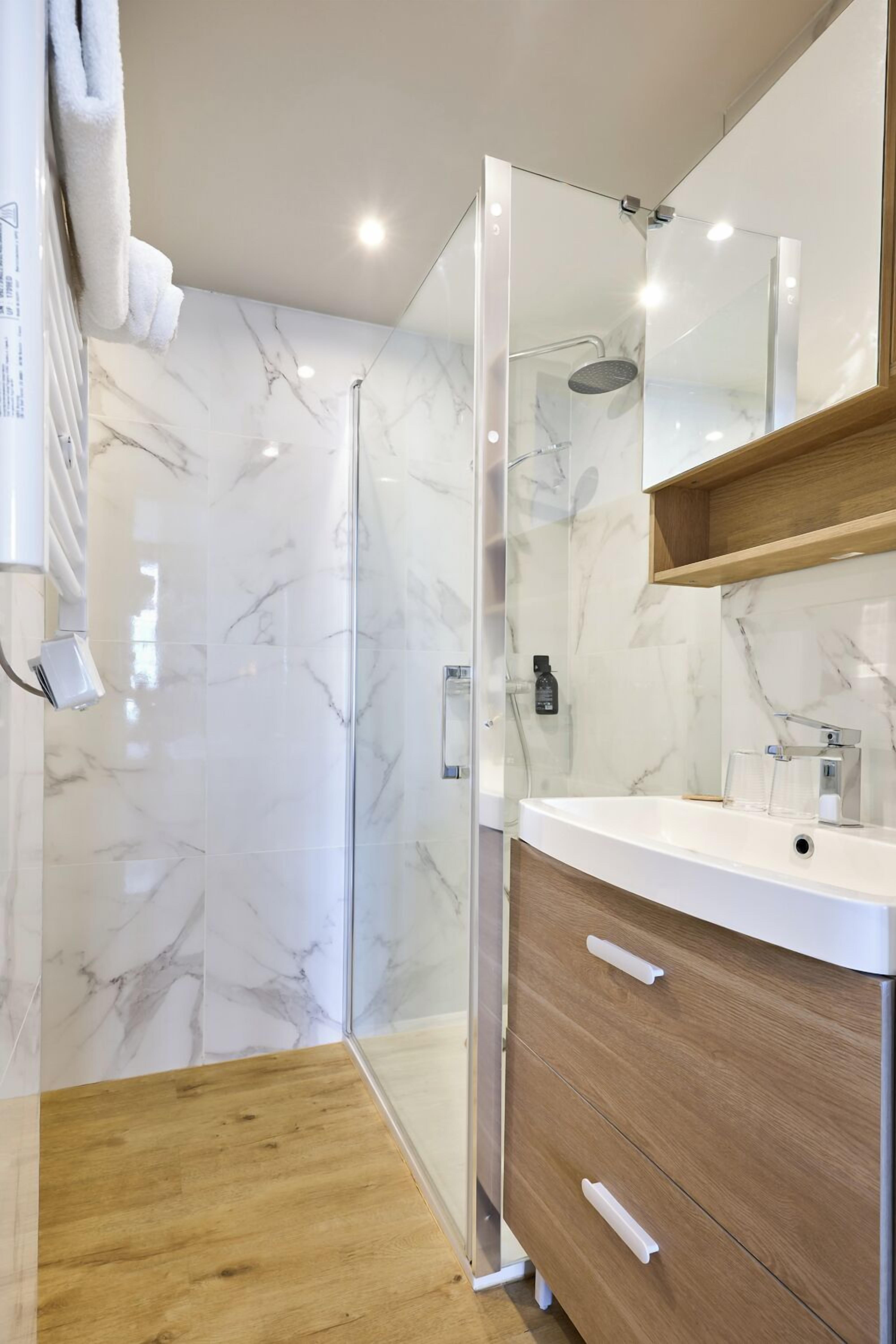 A modern bathroom with a glass shower enclosure, marble walls, a wooden vanity, and a mirror above the sink.