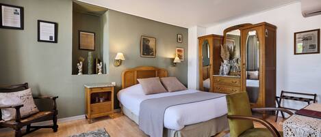 Un dormitorio con cama, mesitas de noche de madera, un sillón verde, un armario de madera y una pequeña mesa con mantel de encaje.