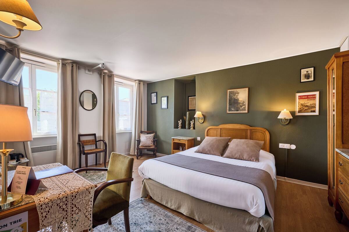 Een hotelkamer met een bed, een bureau met een lamp, een stoel, een tafeltje met een kantkleed en een raam met gordijnen.