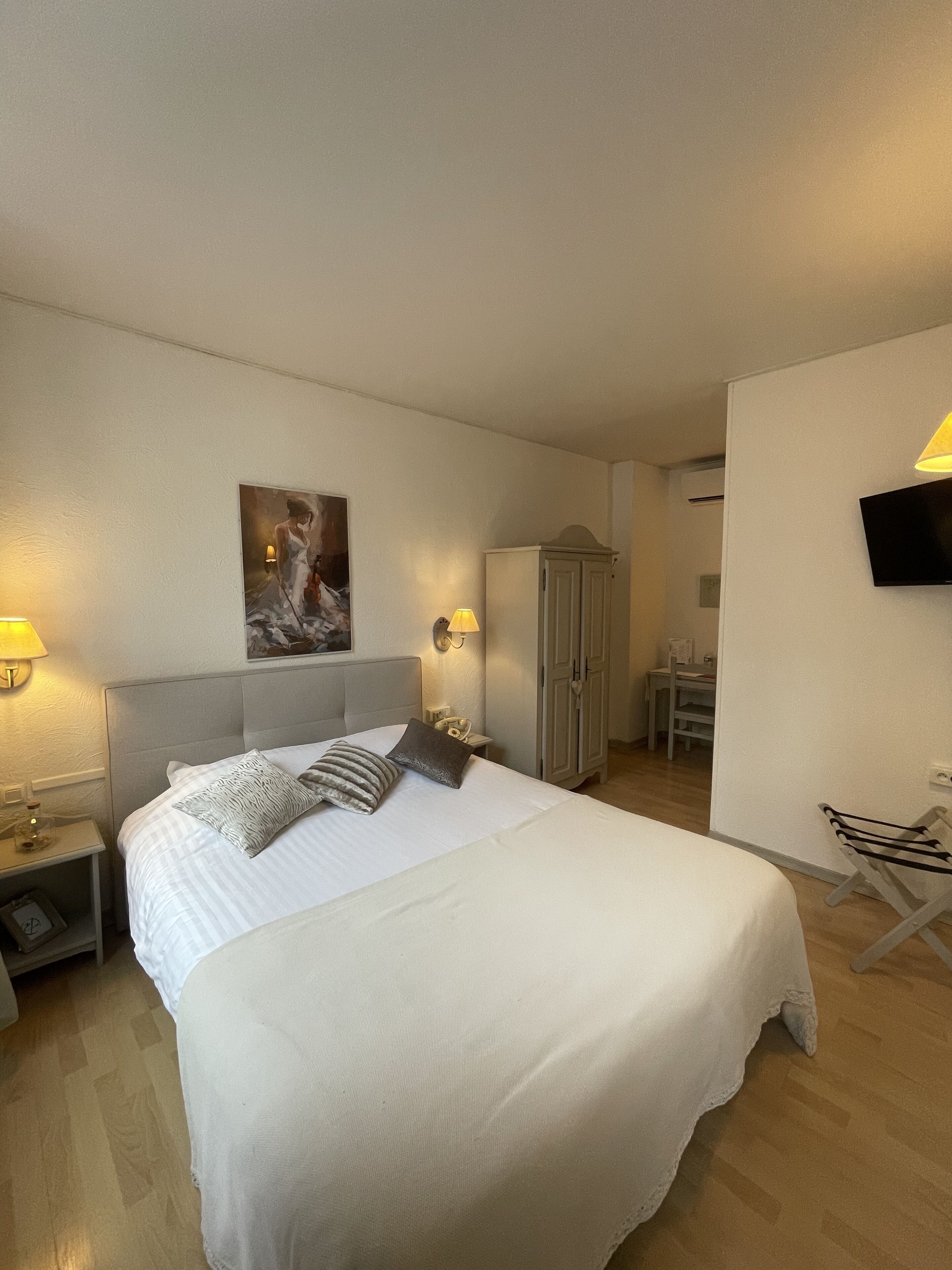 A hotel room with a large bed, bedside lamps, a painting on the wall, a wardrobe, a television, and a small table with a chair.