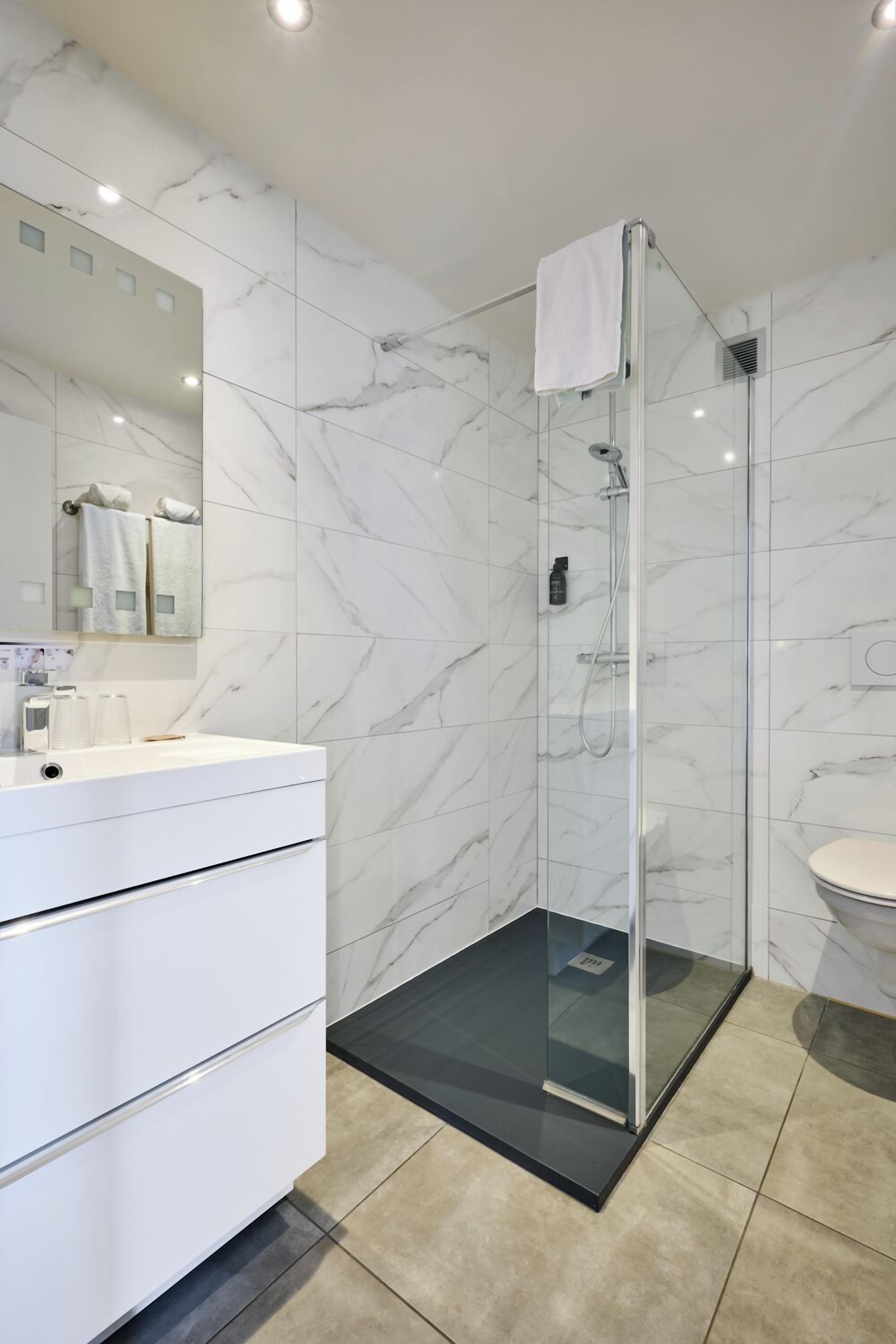 Un baño moderno con ducha envuelta en vidrio, paredes de mármol blanco y tocador blanco con cajones.