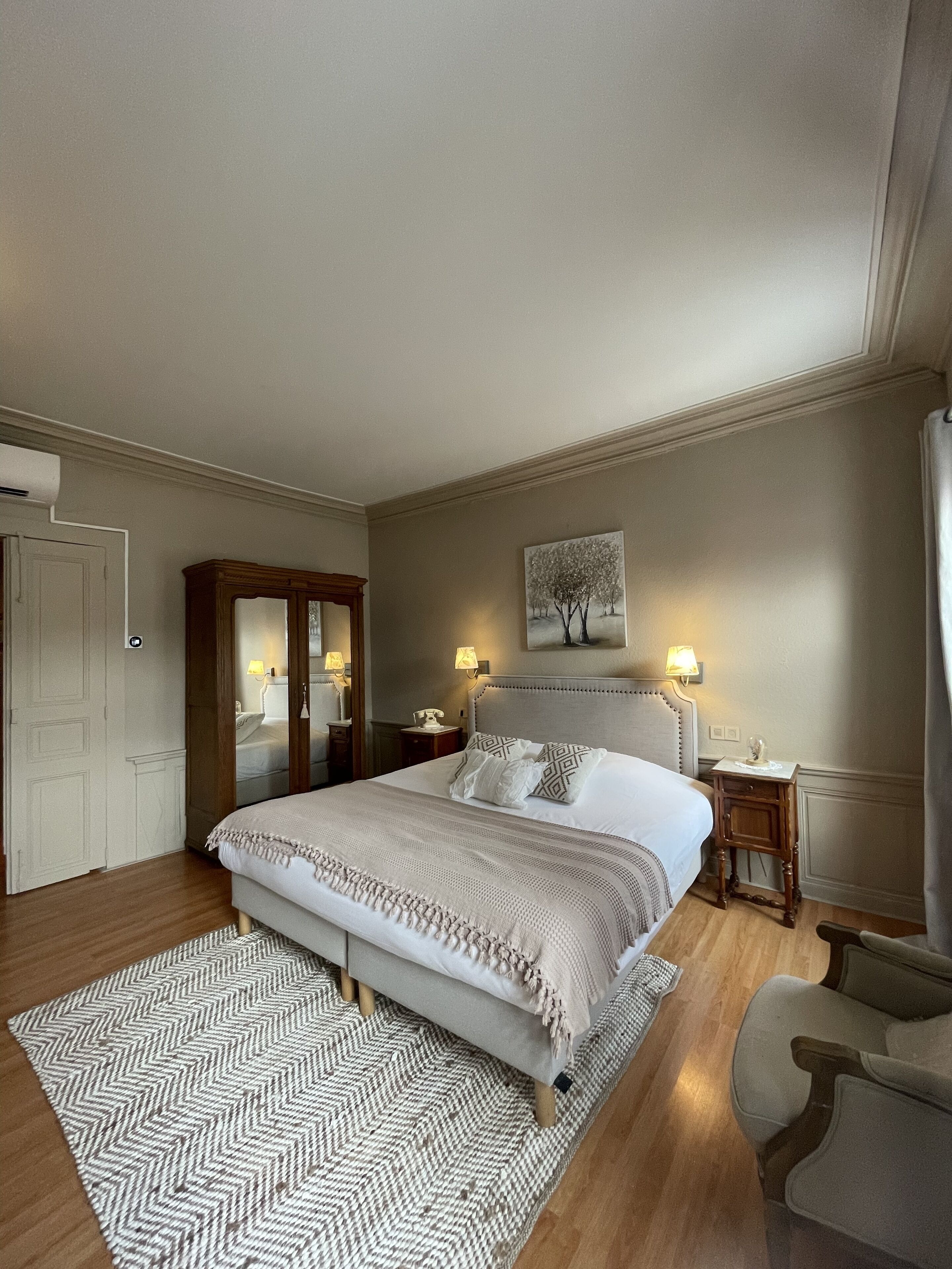 Habitación de hotel con una cama grande, suelo de madera, alfombra estampada en patrón de espiga, mesita de noche con lámpara y un cuadro en la pared.