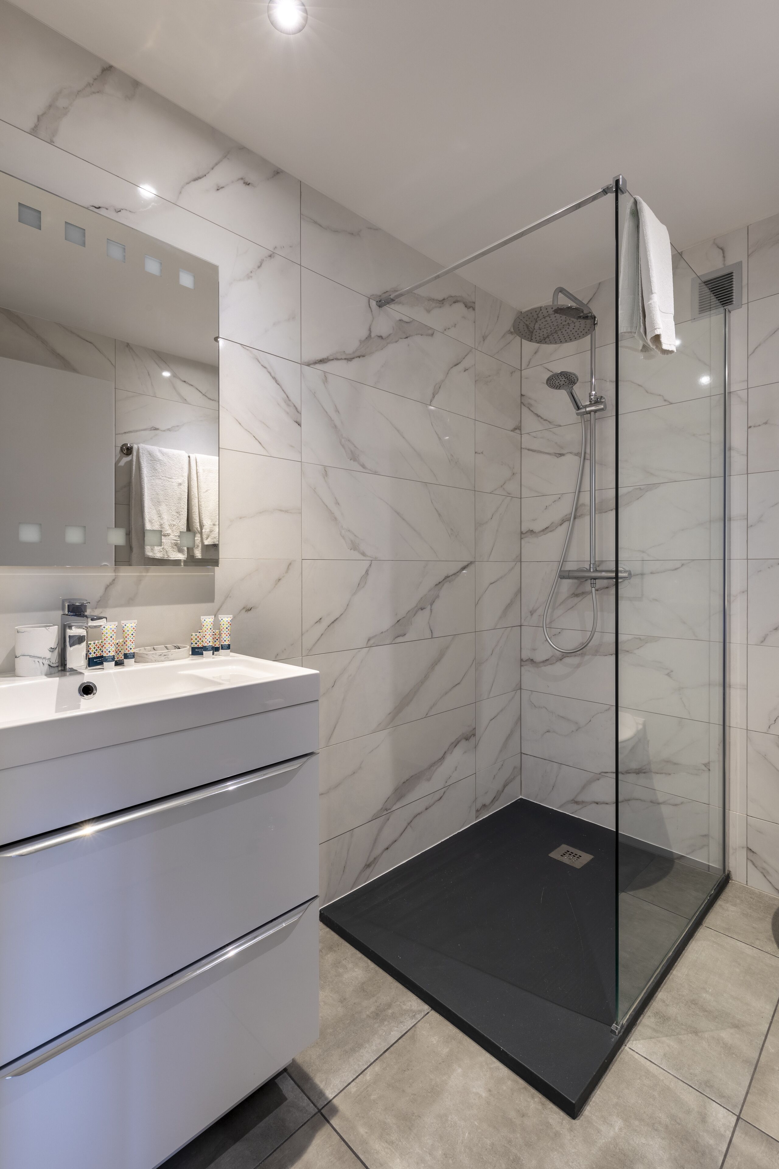 A modern bathroom with a glass-enclosed shower, a white vanity with a mirror, and marble-patterned walls.