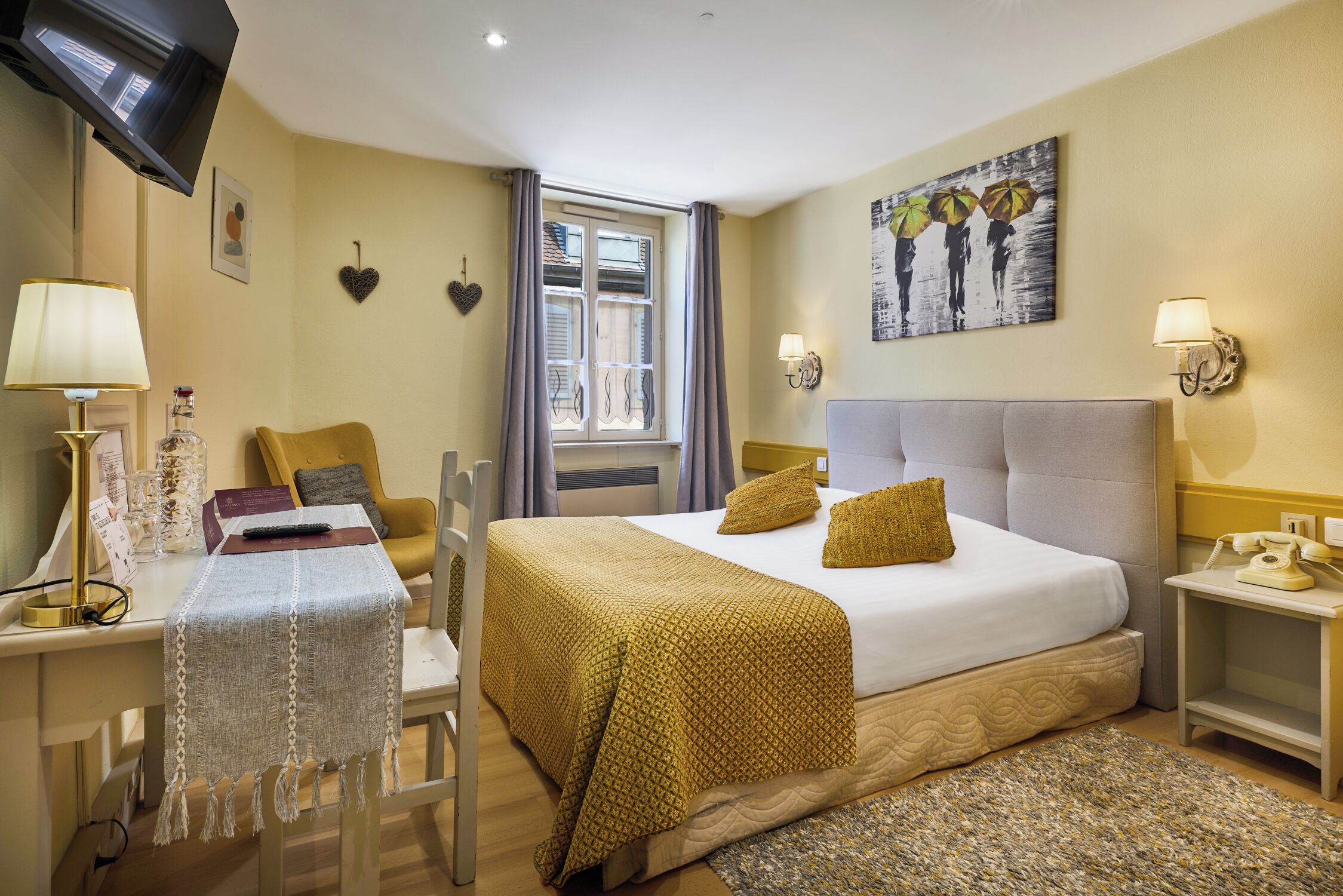 A hotel room with a bed, a desk, a chair, a TV, a window with curtains, a painting on the wall, and a lamp on the nightstand.
