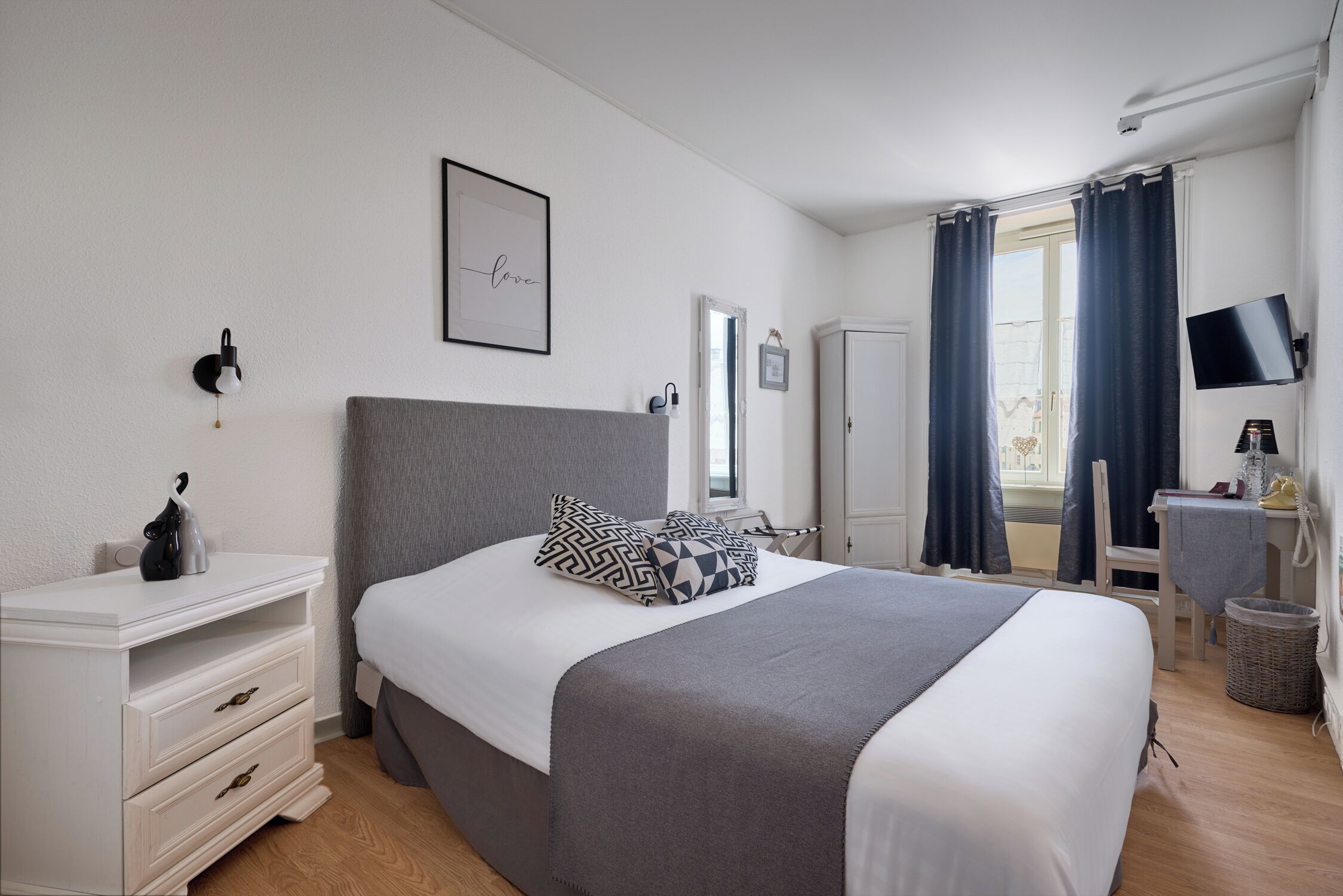 Un dormitorio con cama, mesitas de noche, cómoda, televisor y ventana con cortinas.