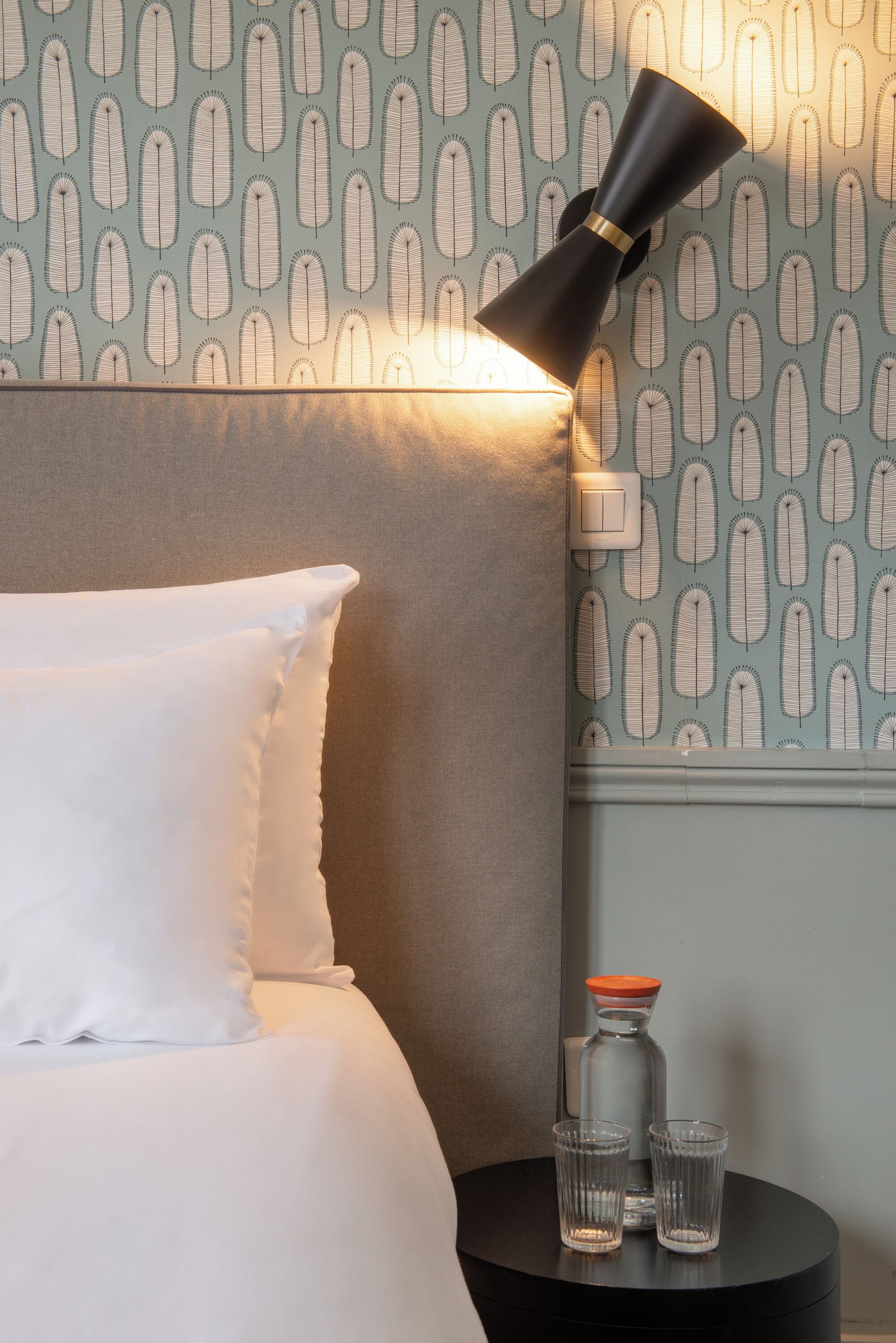 A neatly made bed with white pillows and a black lamp on a bedside table with a patterned wall in the background.