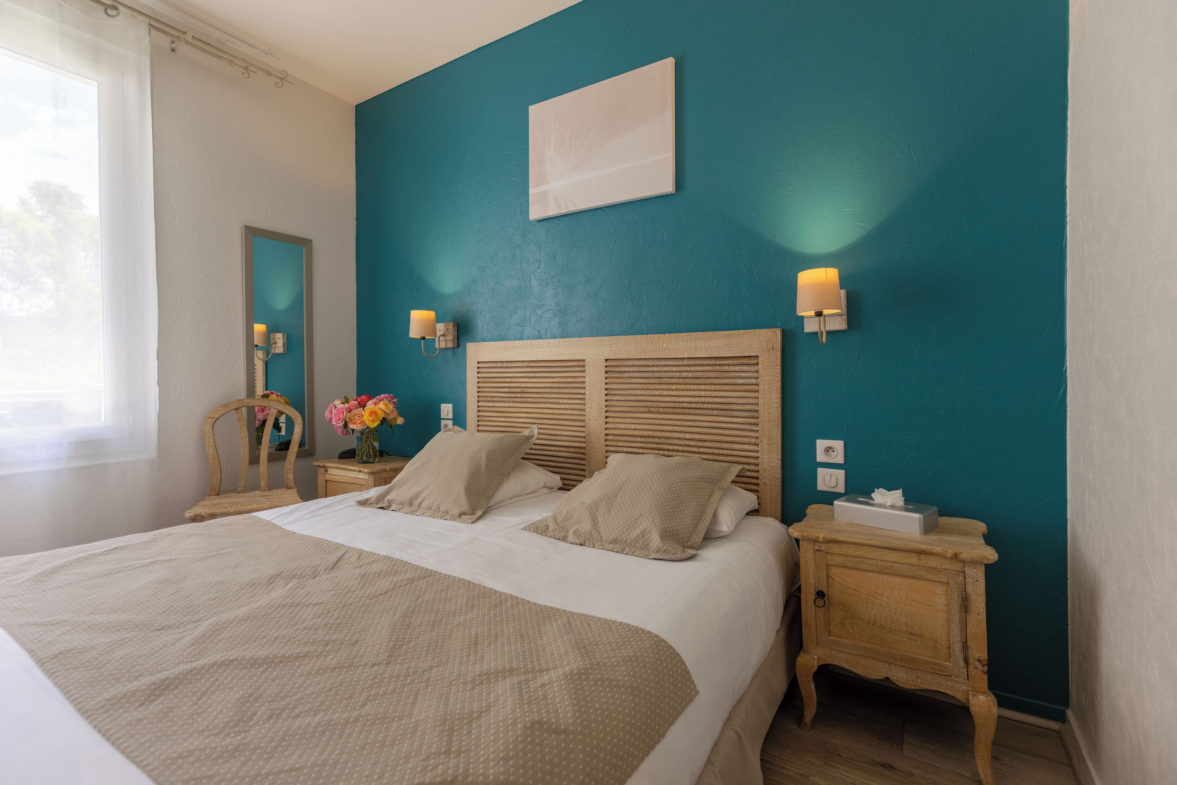 Una cama bien tendida con cabecera de madera, dos mesitas de noche con lámparas, un espejo y un aplique en la pared.