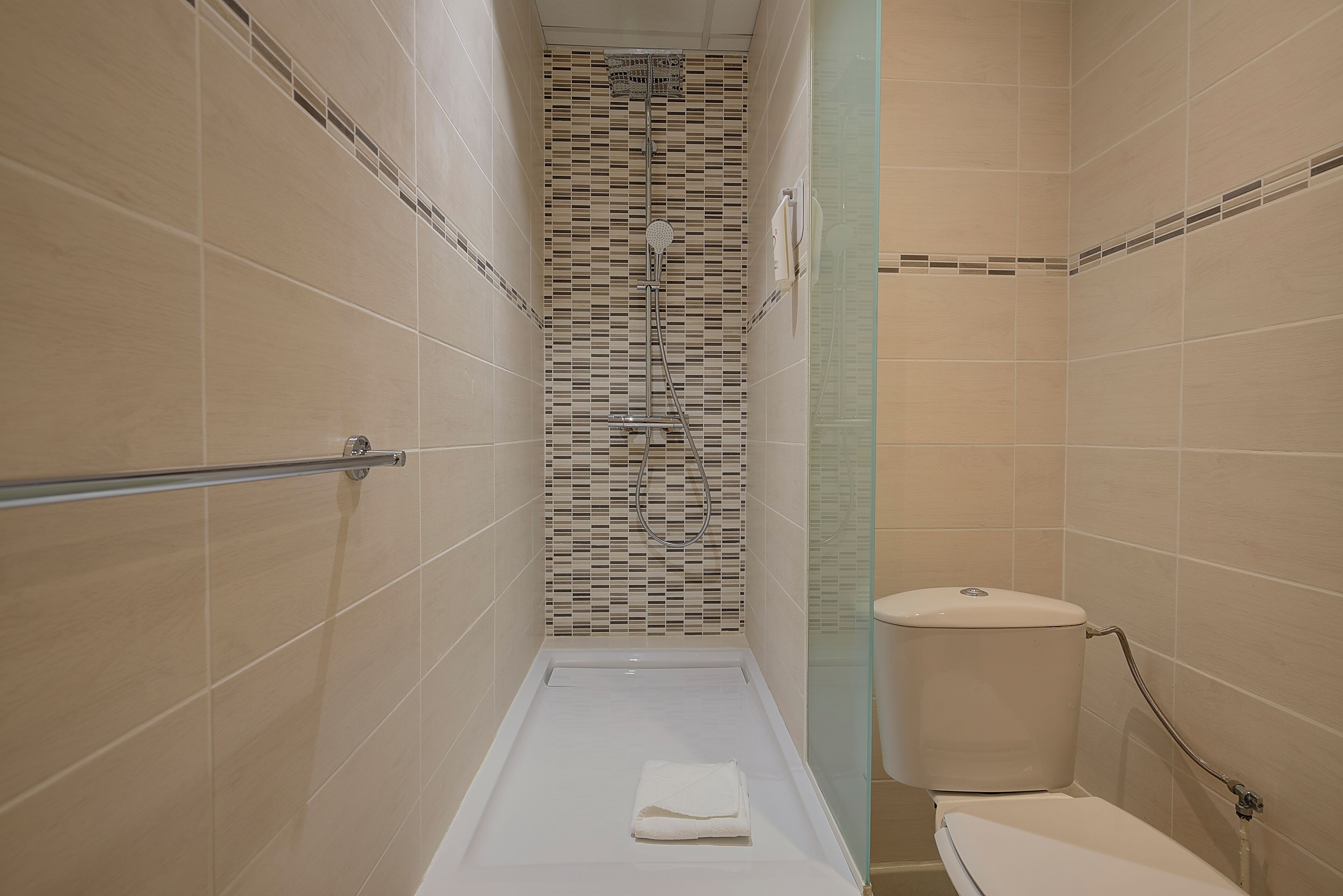 A narrow shower with a glass partition, a toilet, and a towel on the floor.