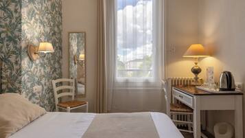 Habitación de hotel con cama, escritorio con lámpara, silla, radiador y ventana con cortinas.