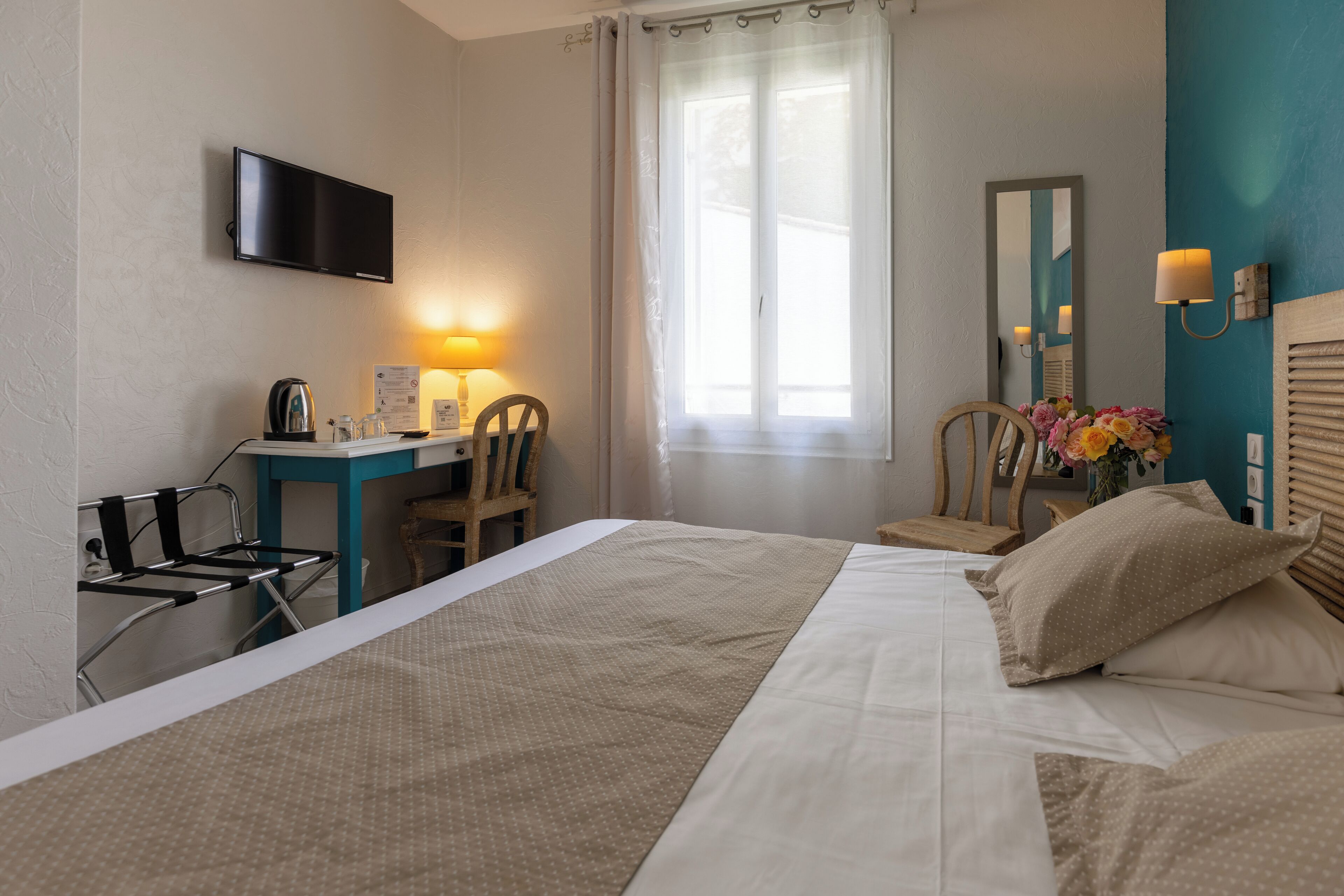 Een hotelkamer met een bed, een bureau met stoel, een televisie, een raam met gordijnen, een spiegel en een wandlamp.