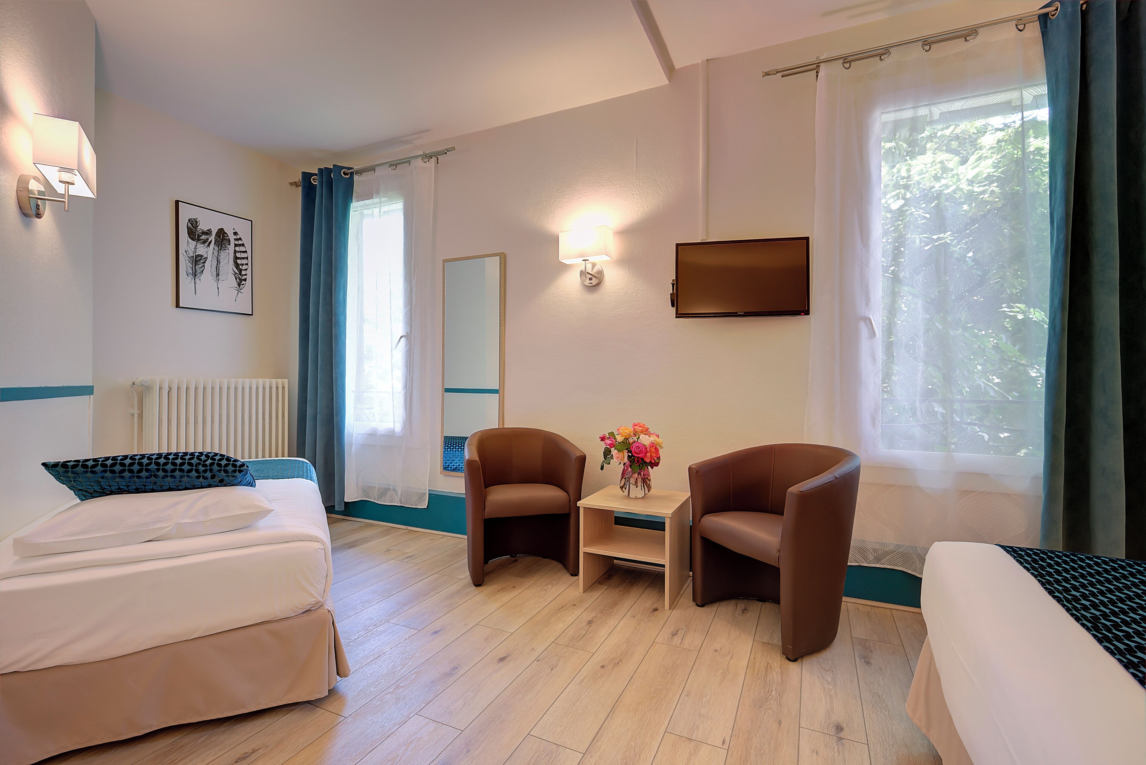 A hotel room with a bed, two armchairs, a small table with flowers, a TV, and a window with curtains.