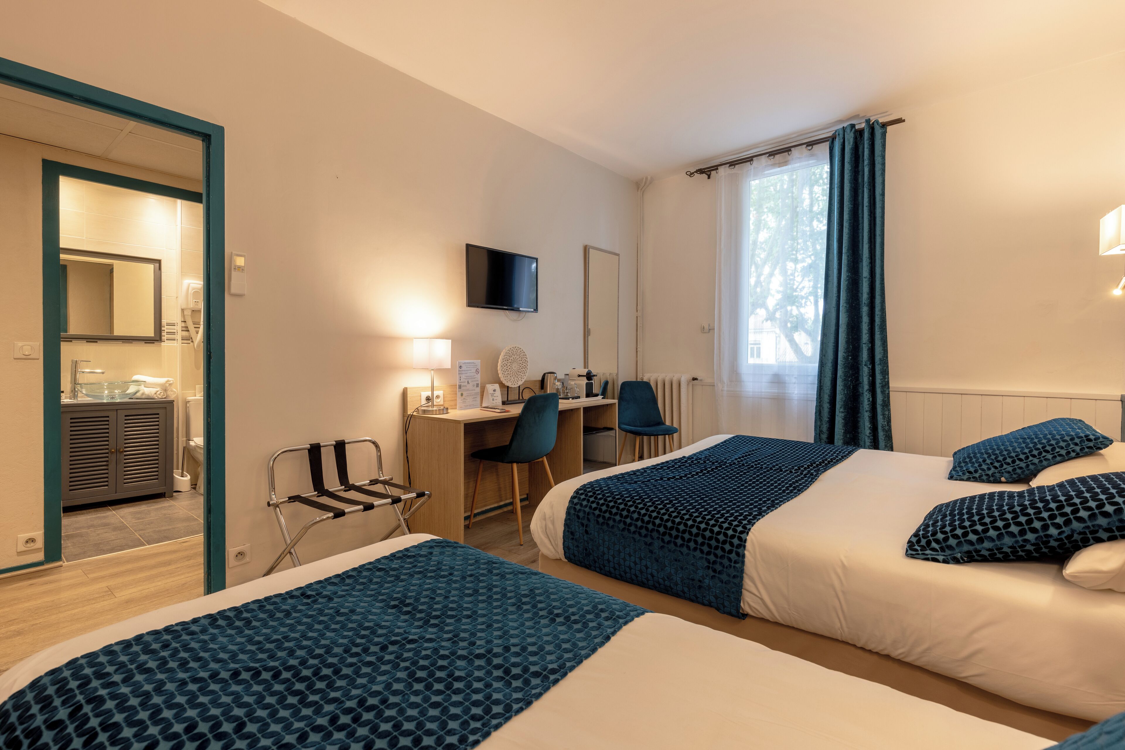 A hotel room with two beds, a desk, a chair, a TV, and a bathroom visible through an open door.