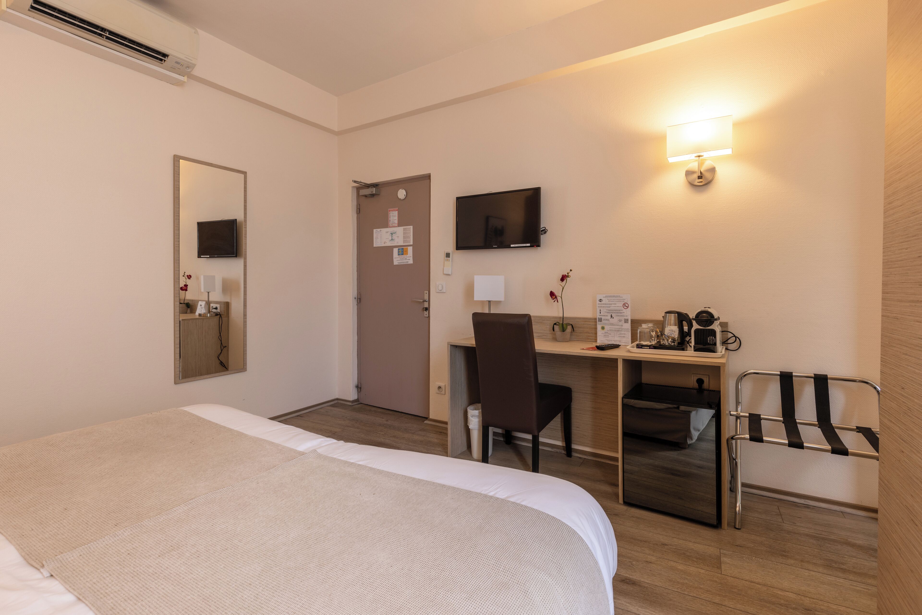 A hotel room with a bed, desk, chair, TV, and a small table with a lamp and a plant.