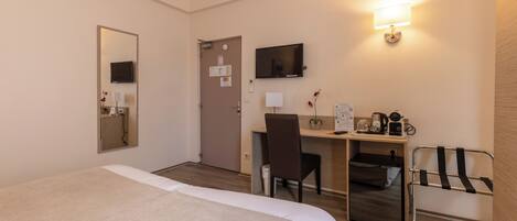 A hotel room with a bed, desk, chair, TV, and a small table with a lamp and a plant.