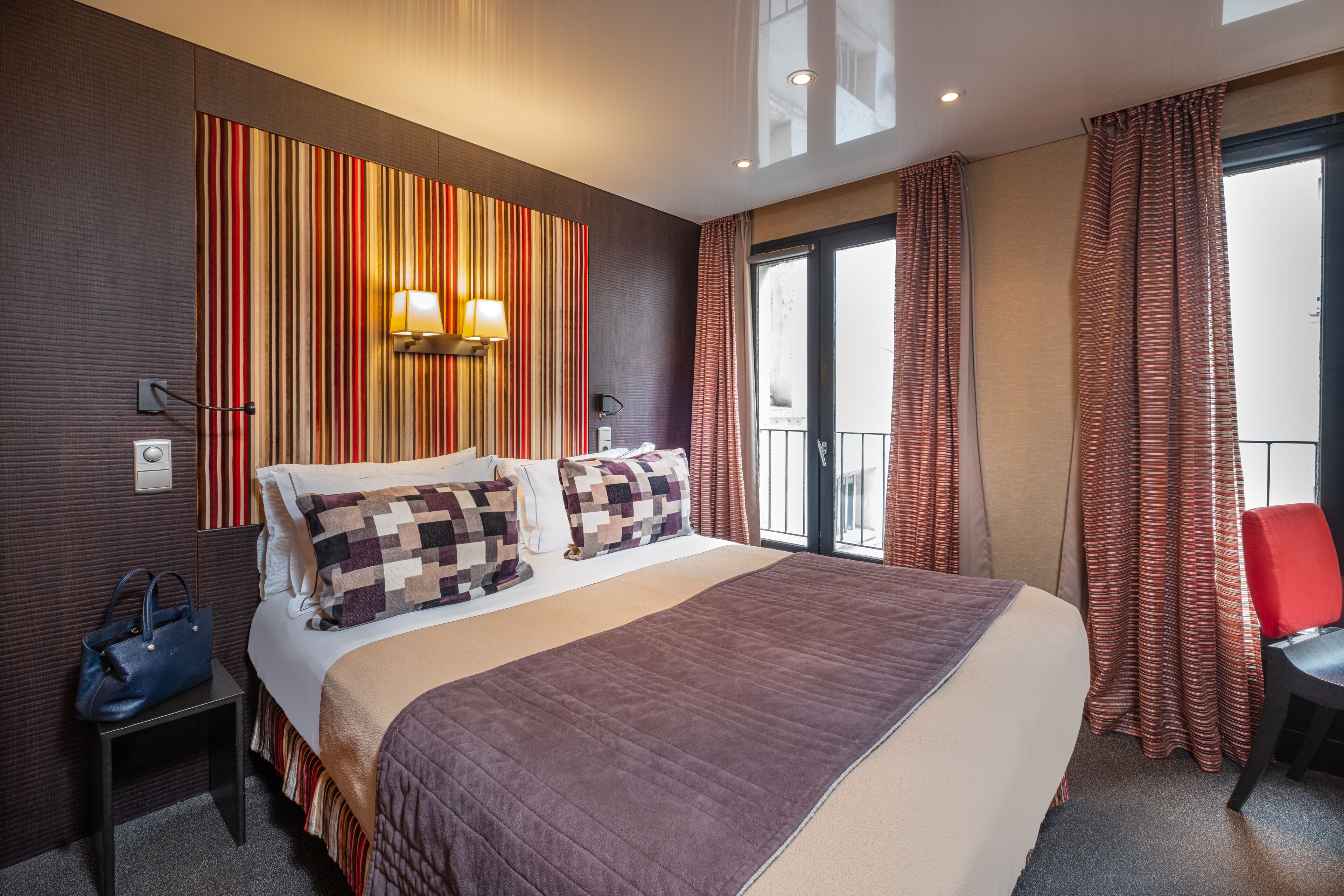 A hotel room with a large bed, a bedside table with a handbag, a wall-mounted lamp, and a view of the outside through a window with curtains.