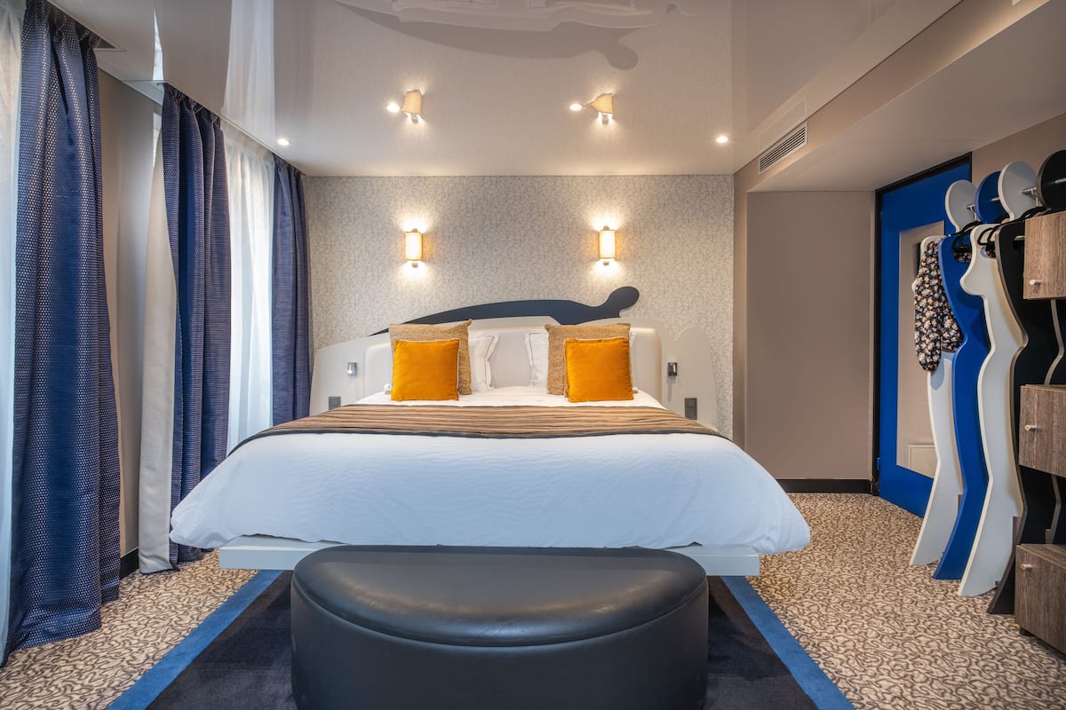 Une chambre d’hôtel moderne dotée d’un grand lit, de deux lampes fixées au mur, d’une tête de lit au design original et d’un mur d’accent bleu.
