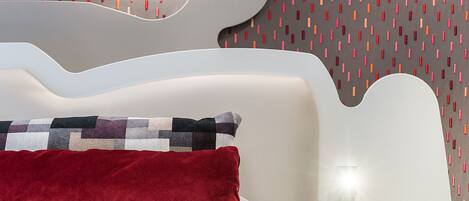Un dormitorio moderno con una cama blanca, un cojín rojo y una pared con estampado.