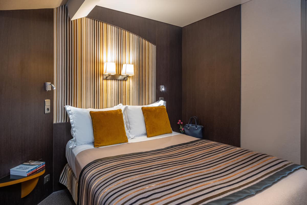Une chambre d’hôtel avec un grand lit, une tête de lit à rayures, deux lampes de chevet, une table de chevet avec des livres et un sac bleu posé sur le sol.