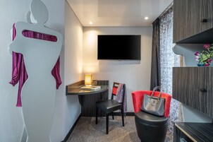 Ein modernes Hotelzimmer mit einem flachbildfernseher an der Wand, einem kleinen Schreibtisch mit Lampe, einem Sessel und einer roten Handtasche auf einem schwarzen Pouf.