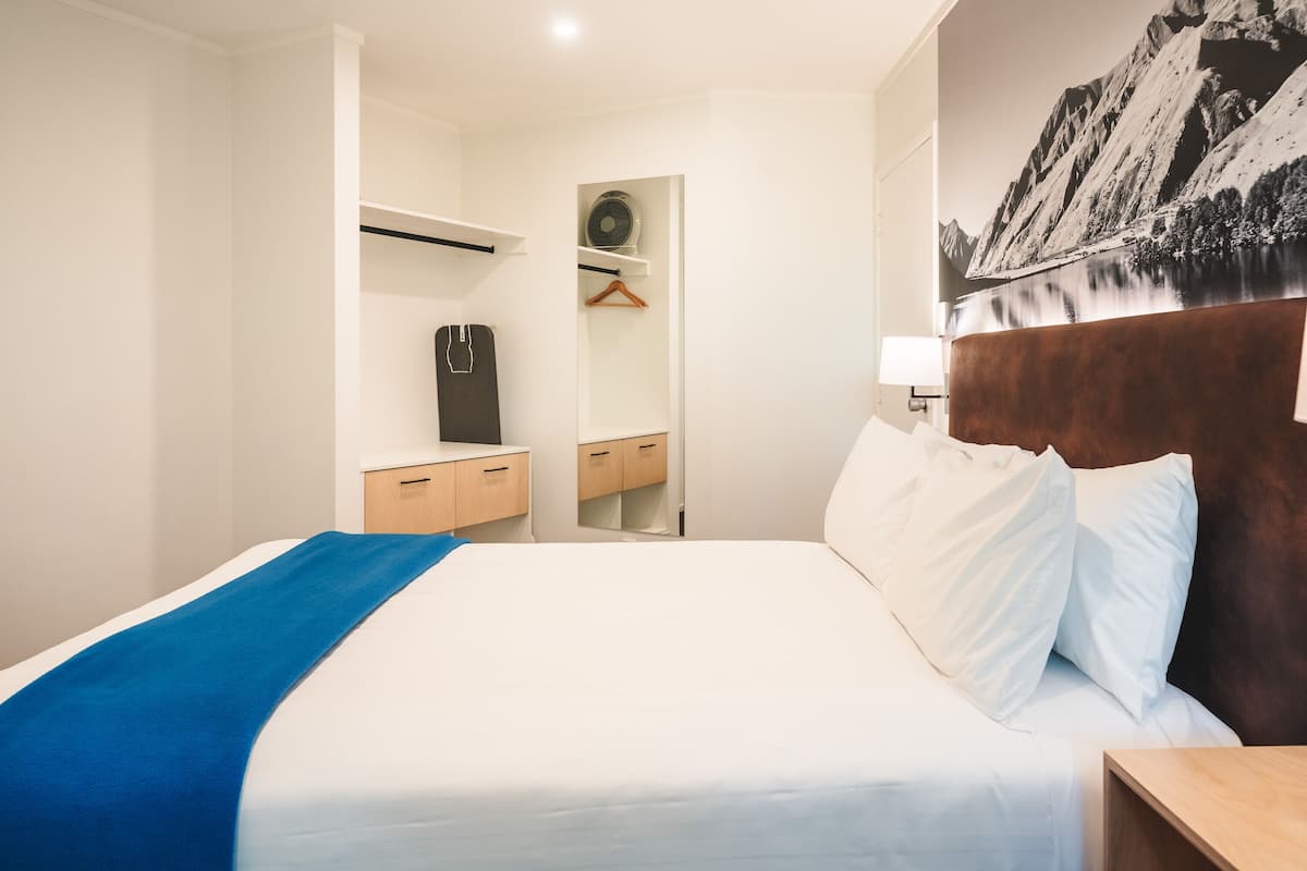 Una habitación de hotel con una cama bien tendida, una mesita de noche de madera, un aparador y un espejo.