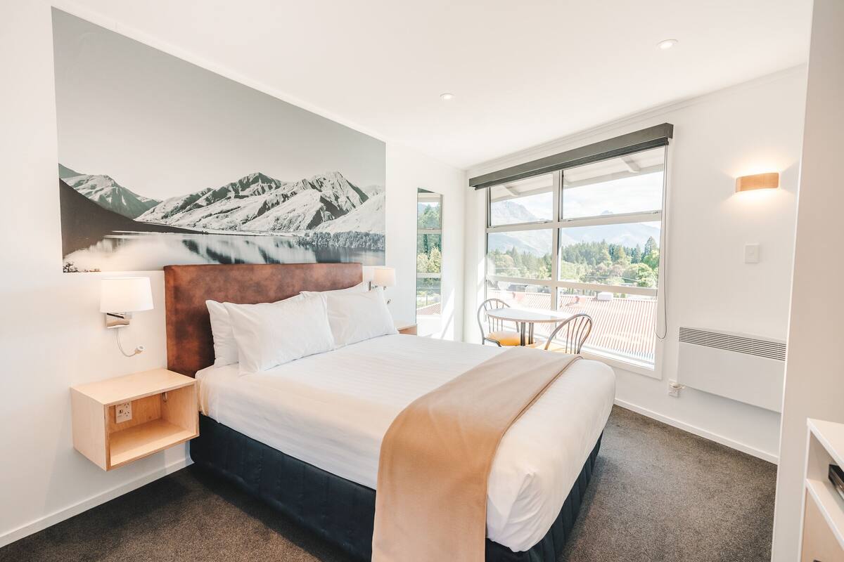 Una habitación de hotel moderna con una cama grande, vistas a las montañas a través de la ventana, una pequeña mesa con sillas y una lámpara fijada a la pared.