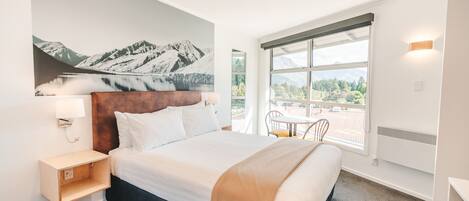 Een moderne hotelkamer met een groot bed, uitzicht op bergen door het raam, een kleine tafel met stoelen en een wandlamp.
