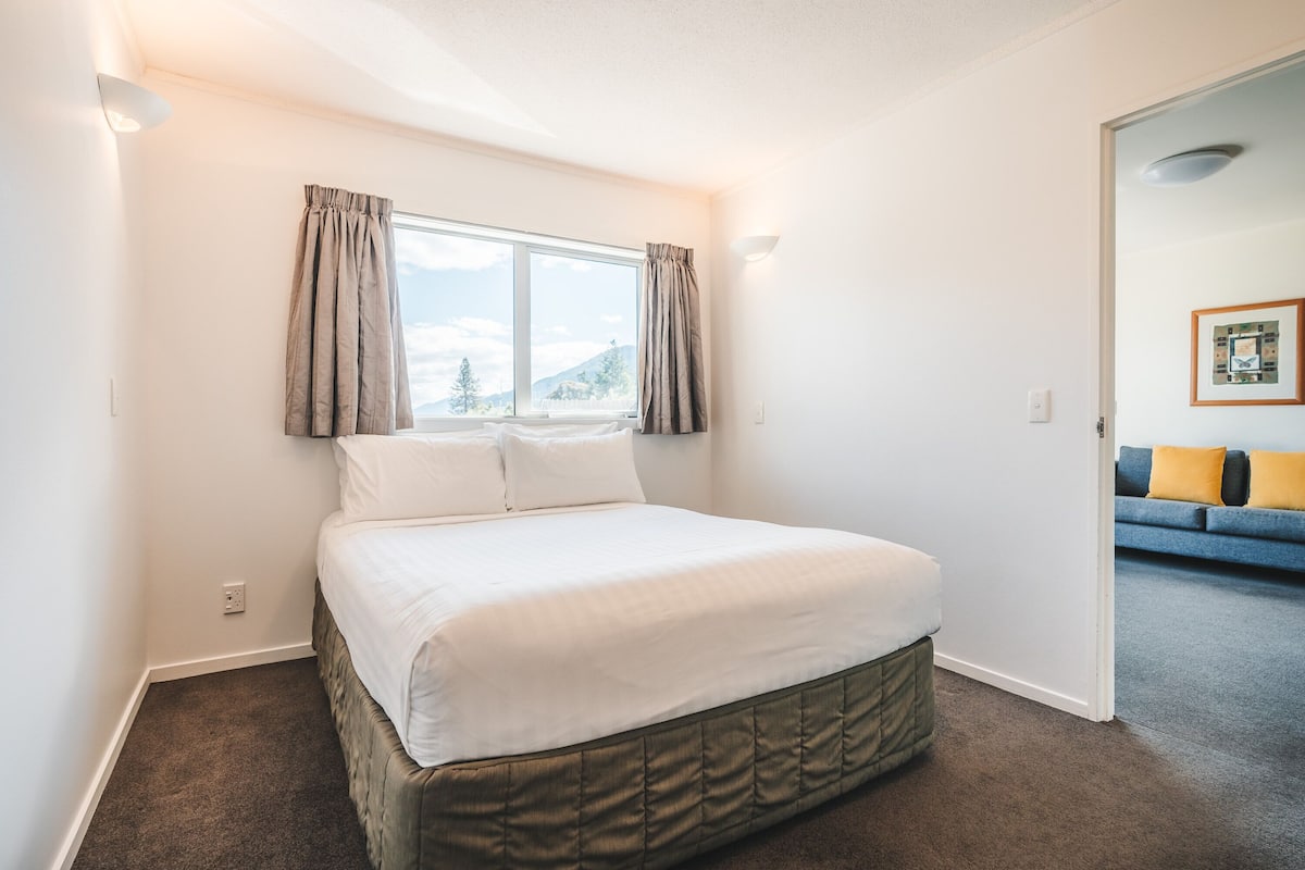 Un dormitorio con cama, ventana con cortinas, luz empotrada en la pared y puerta que da a una sala de estar con sofá.
