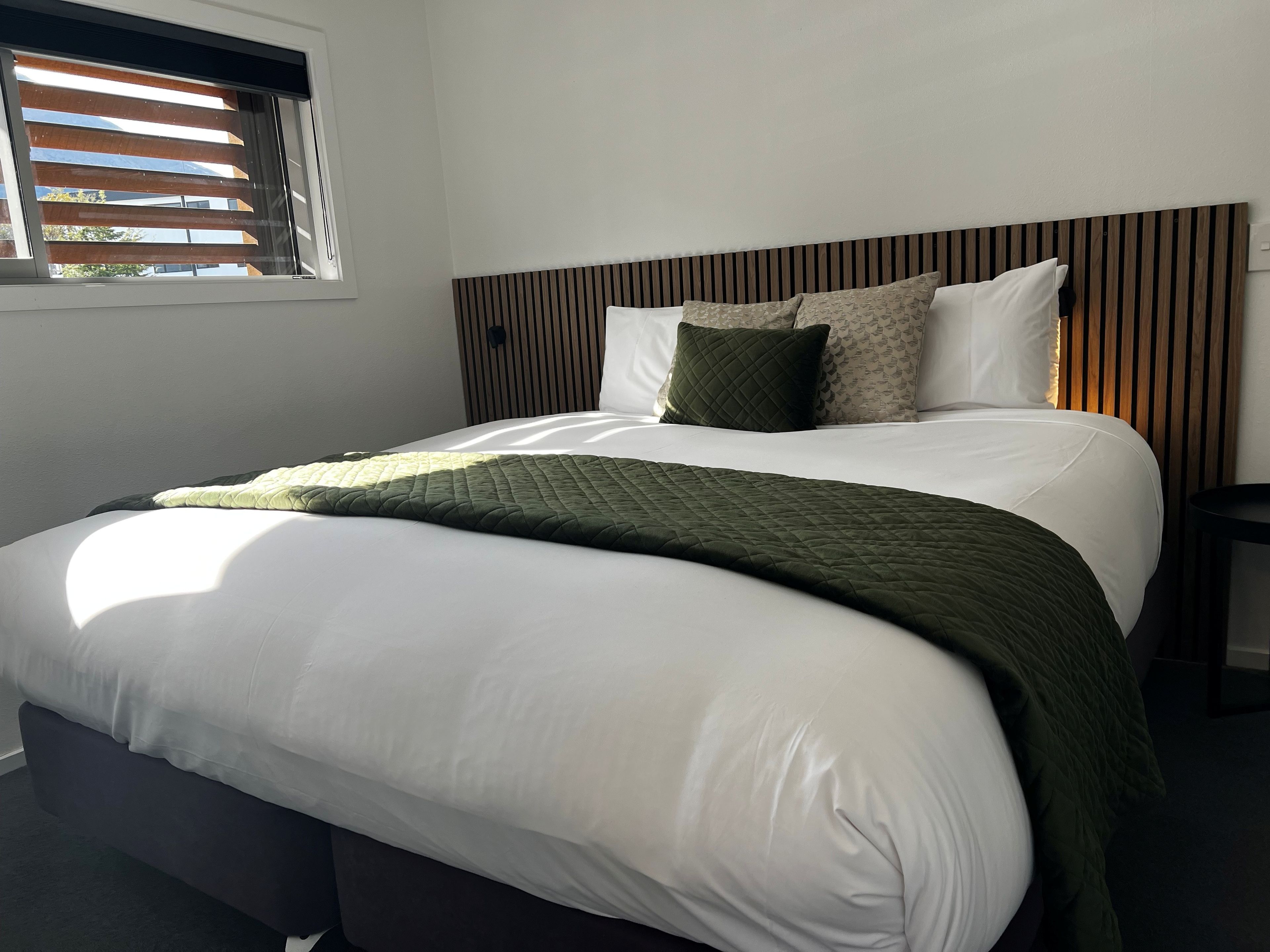 Ein ordentlich bezogenes Bett mit einer gesteppten, grünen Decke, weißer Bettwäsche und einem hölzernen Kopfteil.