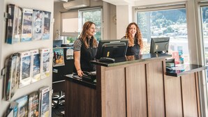 A reception desk with a wooden counter, a computer, and a ViewSonic monitor. There are brochures on the wall and a window with a view of trees. - Blue Peaks Lodge (Queenstown)